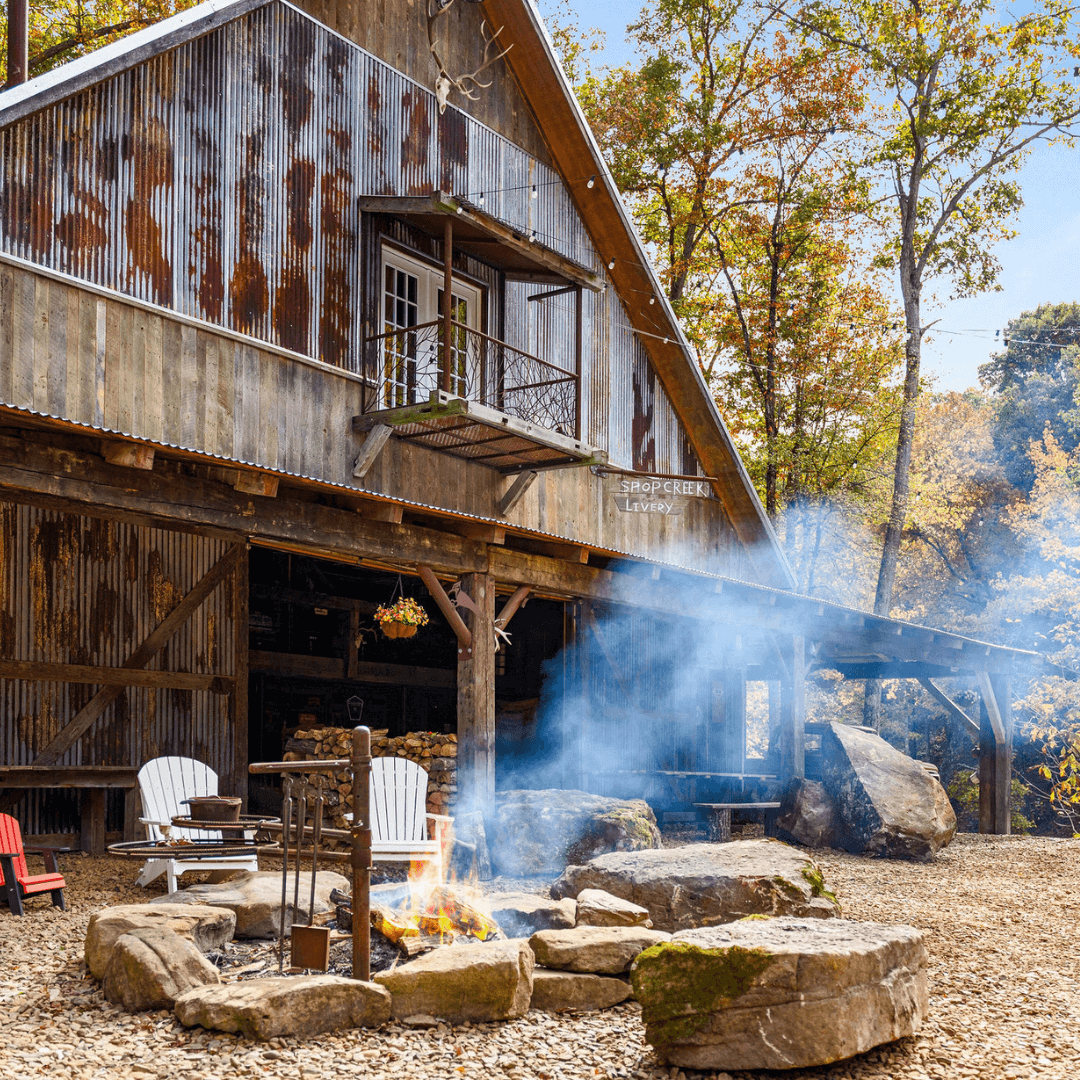 Rustic cabin with corrugated metal siding and a small balcony, surrounded by autumn trees. In the foreground, a stone fire pit with seating, and smoke rising from the fire. A sign on the cabin reads 'Shopcreek Levey.'