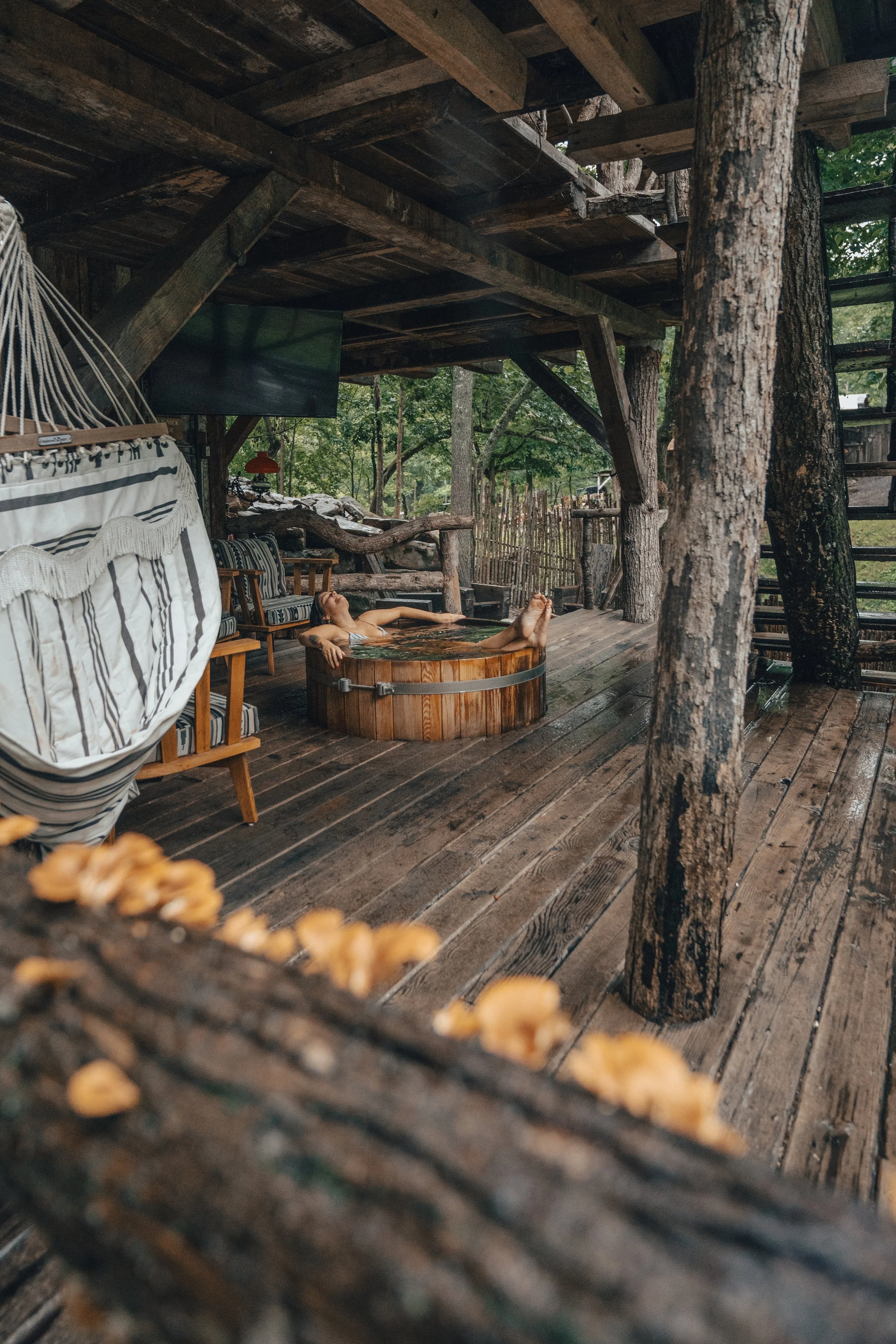 A woman relaxing in a wooden hot tub outdoors, surrounded by trees and rustic wooden structures.