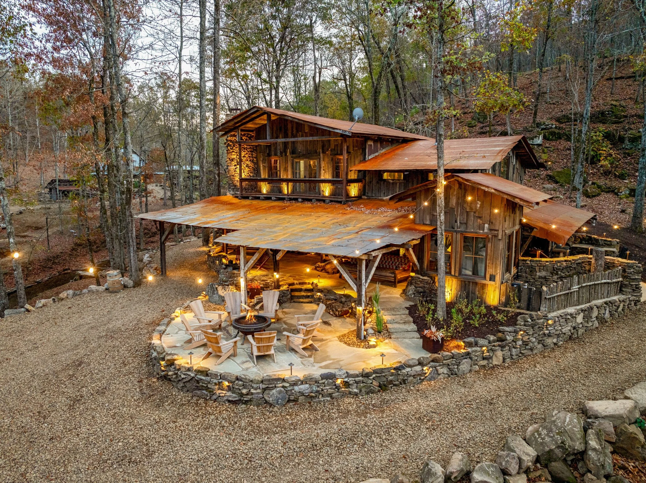 Rustic cabin with a wraparound porch surrounded by woods, featuring wooden chairs around a fire pit and warm ambient lighting.