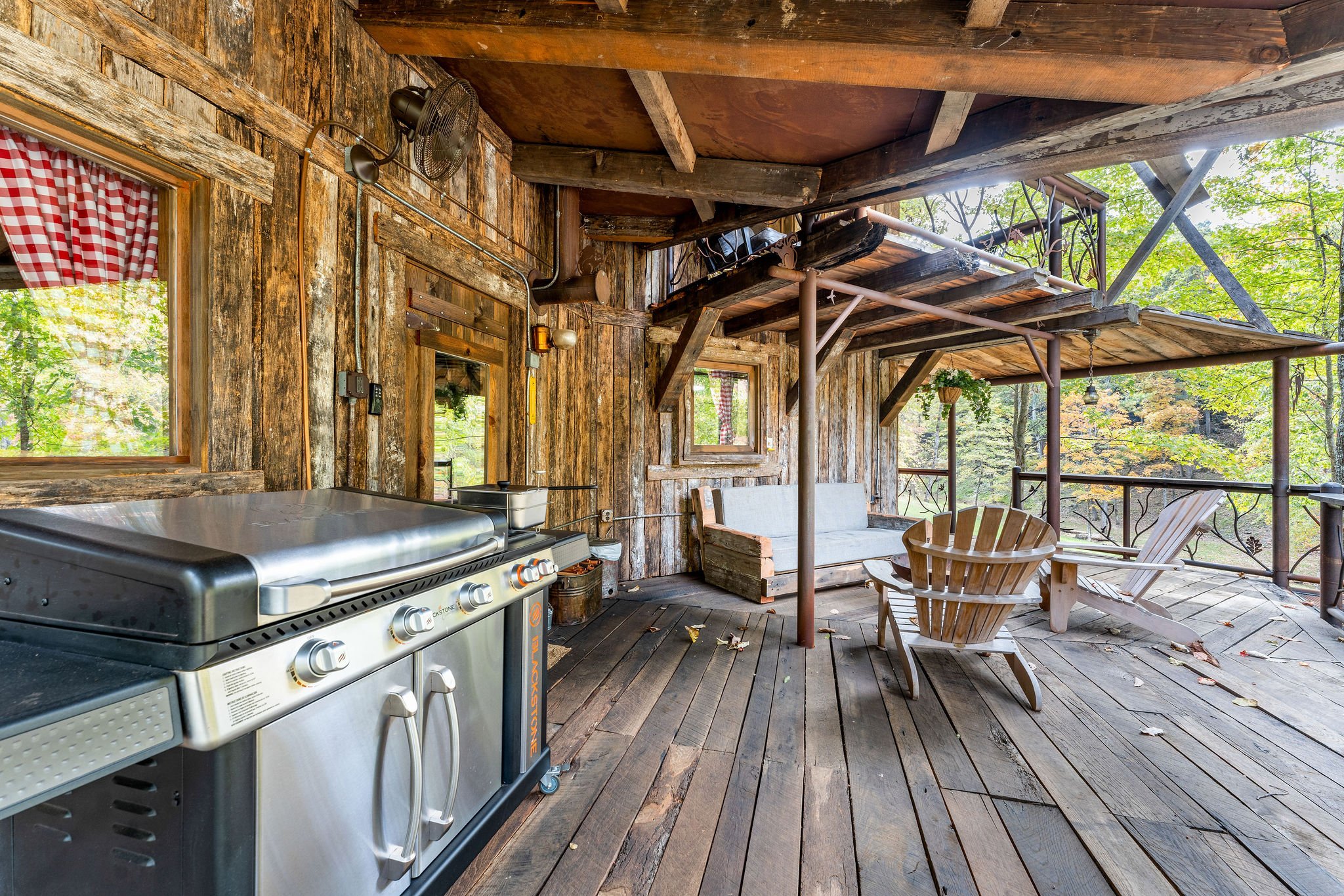 Rustic wooden patio with a grill, Adirondack chairs, a wooden bench, exposed beams, and trees visible in the background.