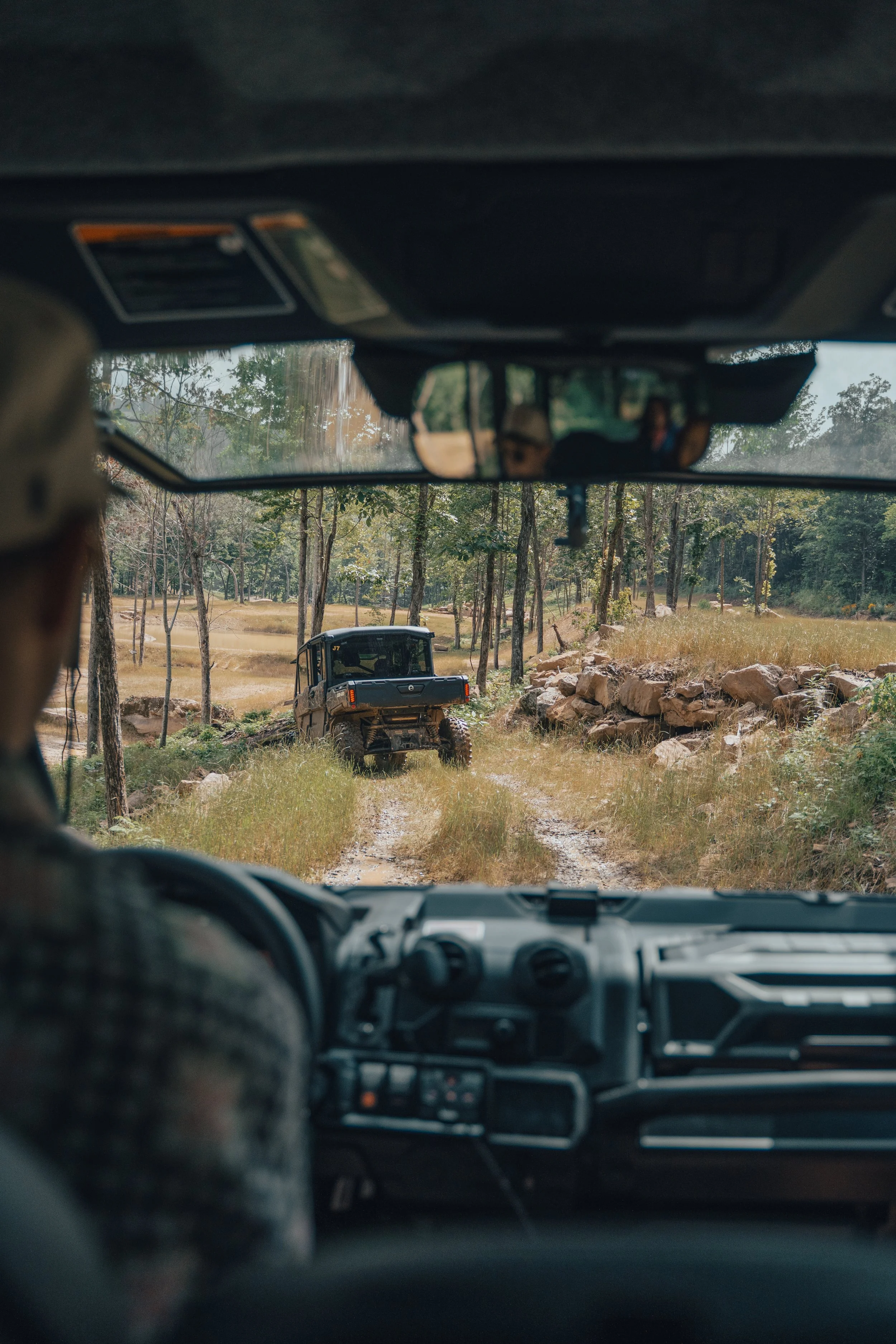View from inside a vehicle looking through the front windshield at a dirt trail in a wooded area with an off-road utility vehicle ahead, surrounded by trees and rocks.