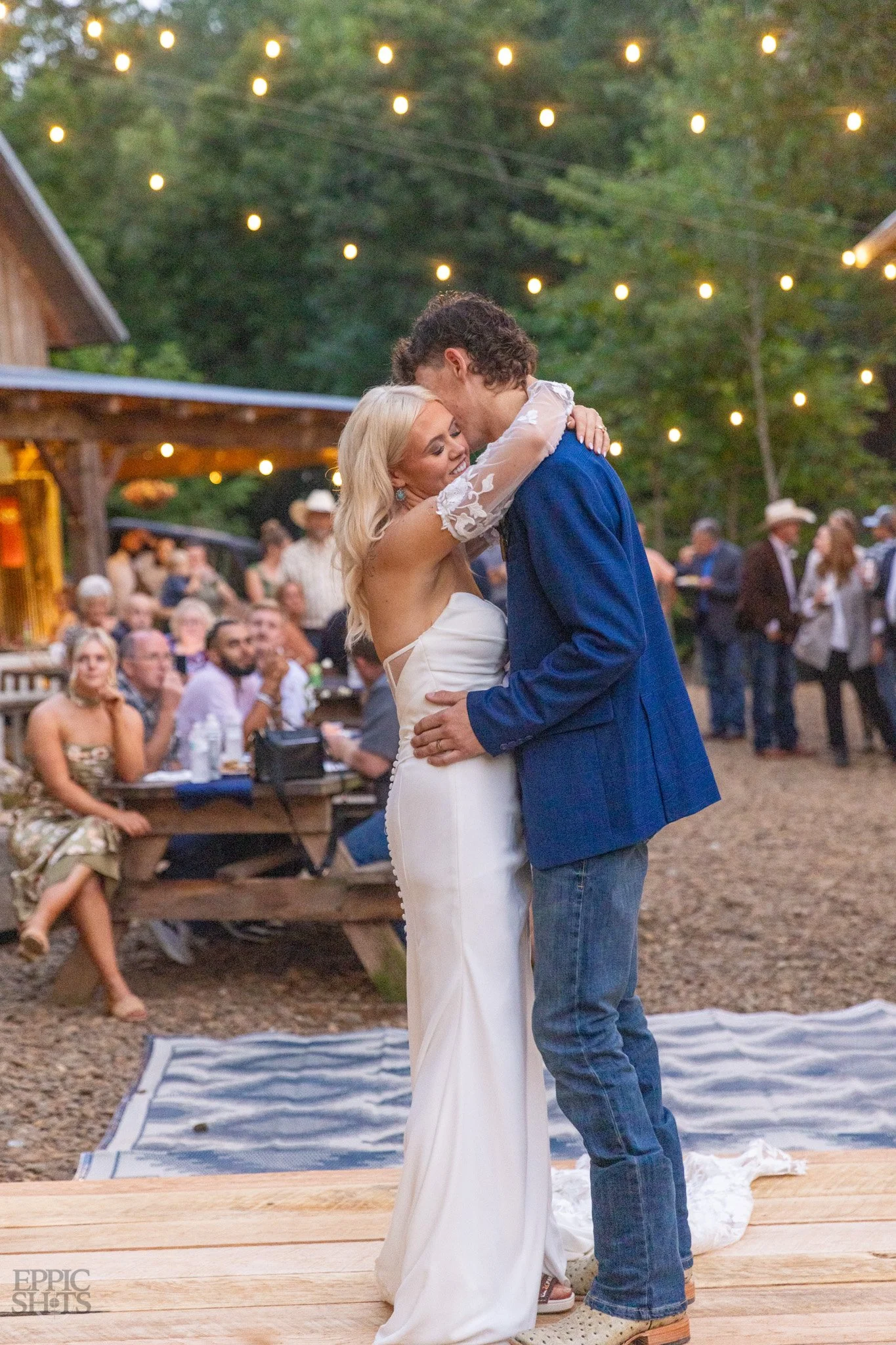 A bride and groom engaged in a dance during an outdoor wedding reception at dusk, under string lights, with guests sitting and standing around in the background.