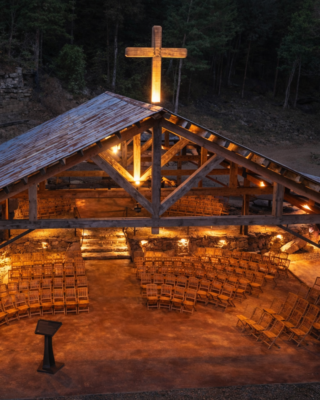 Outdoor covered chapel with wooden structure, rows of chairs arranged for a service, illuminated by warm lighting, with a cross mounted on a hill in the background surrounded by trees.