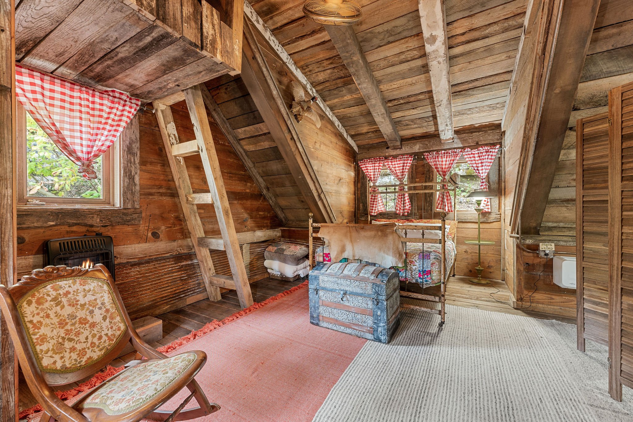 Rustic attic bedroom with wooden walls, red checkered curtains, vintage iron bed, antique chest, patterned rug, and floral upholstered chair.
