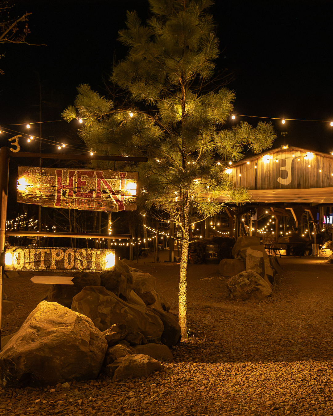 Night scene with a tree illuminated by string lights, rocks on the ground, and a building with string lights in the background, featuring signs that say 'HEAVEN' and 'BOOTH POST'.