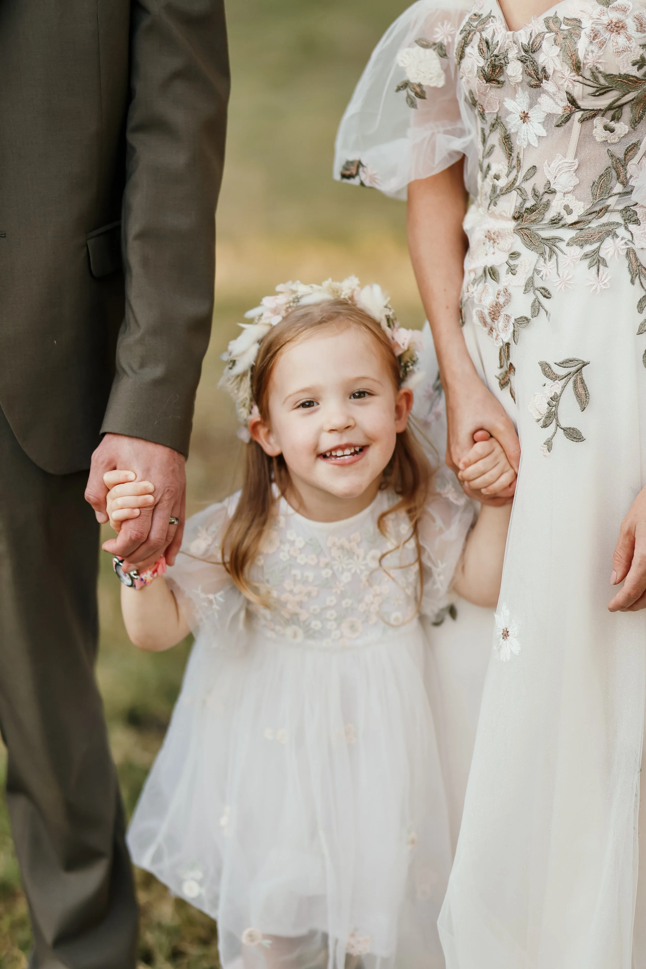 A young girl with red hair, wearing a white dress with floral embroidery and a flower crown, holding hands with two adults during a wedding ceremony.