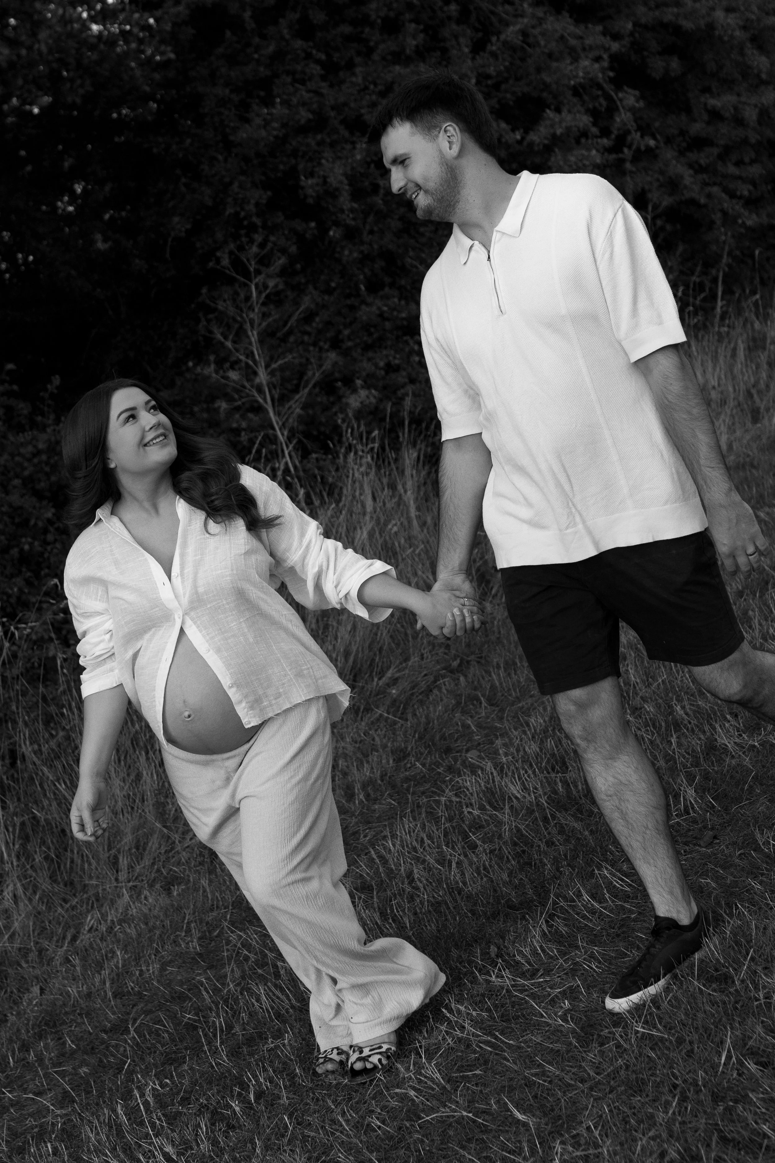 A pregnant woman and a man walk hand in hand outdoors, smiling at each other, with the woman looking up at the man.