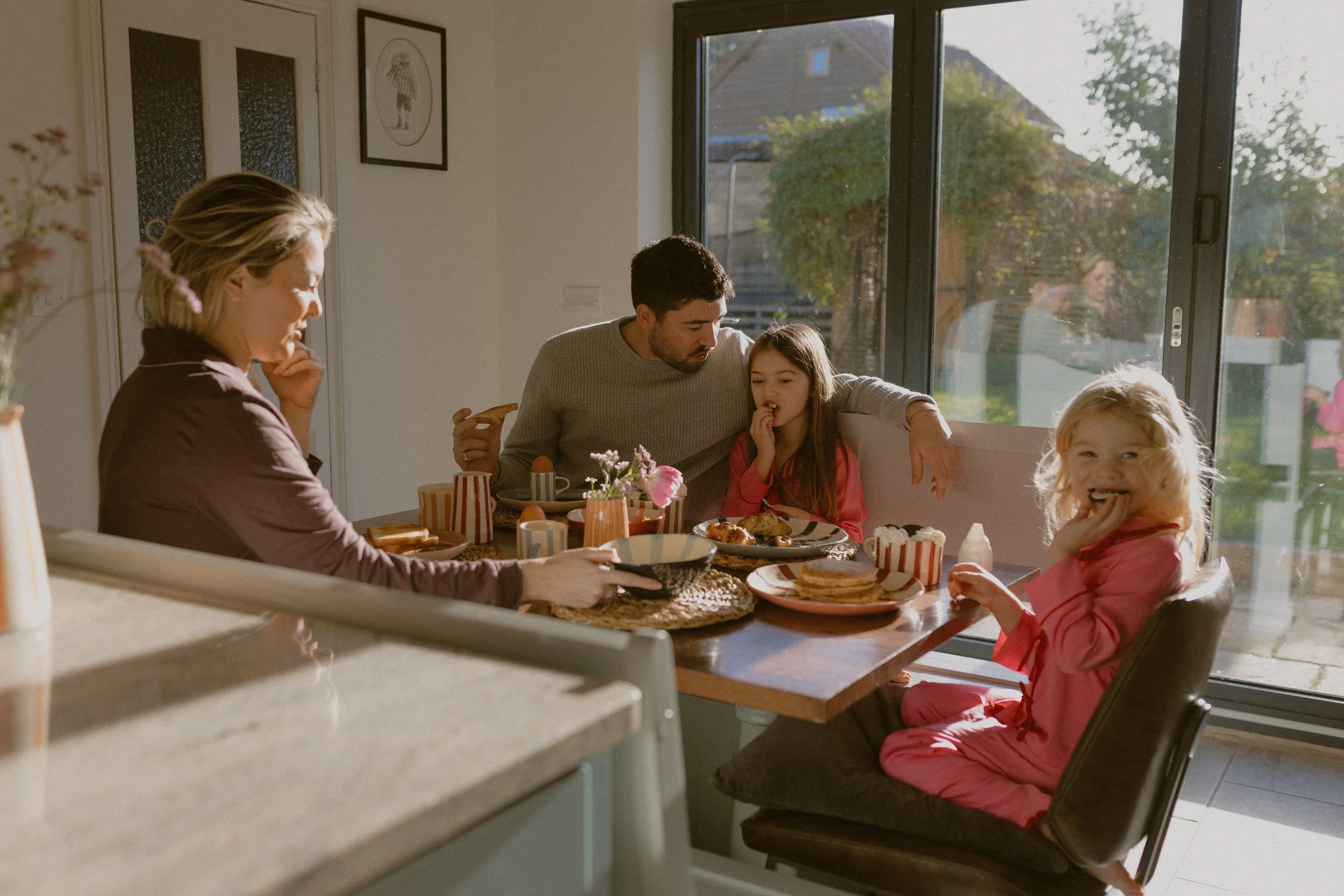 Family having breakfast at the kitchen table, with sunlight coming through a glass door, showing their backyard in the background.