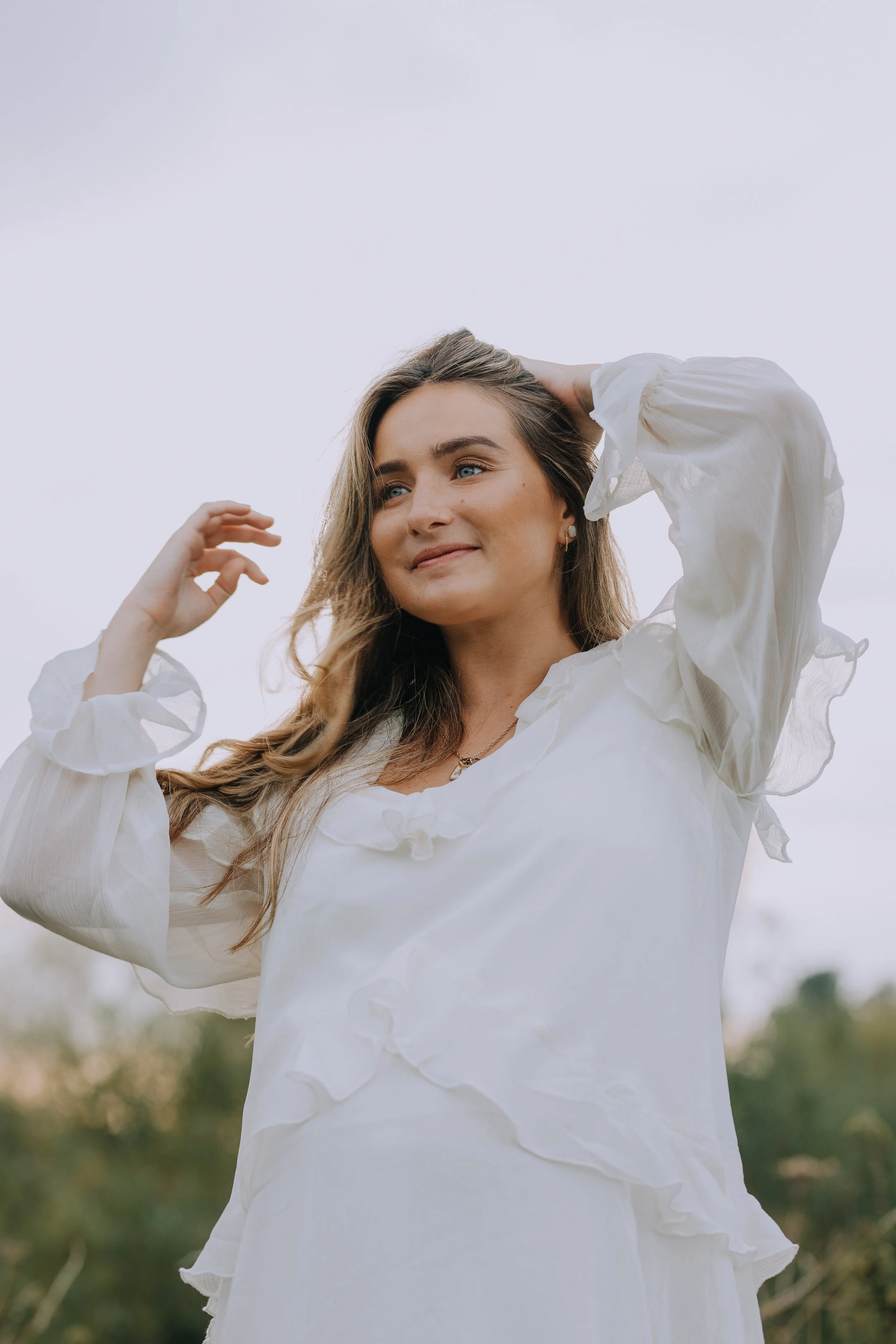 A young woman with light brown hair and blue eyes standing outdoors in a white flowy dress, looking to the side with a gentle smile, against a cloudy sky and blurred trees in the background.
