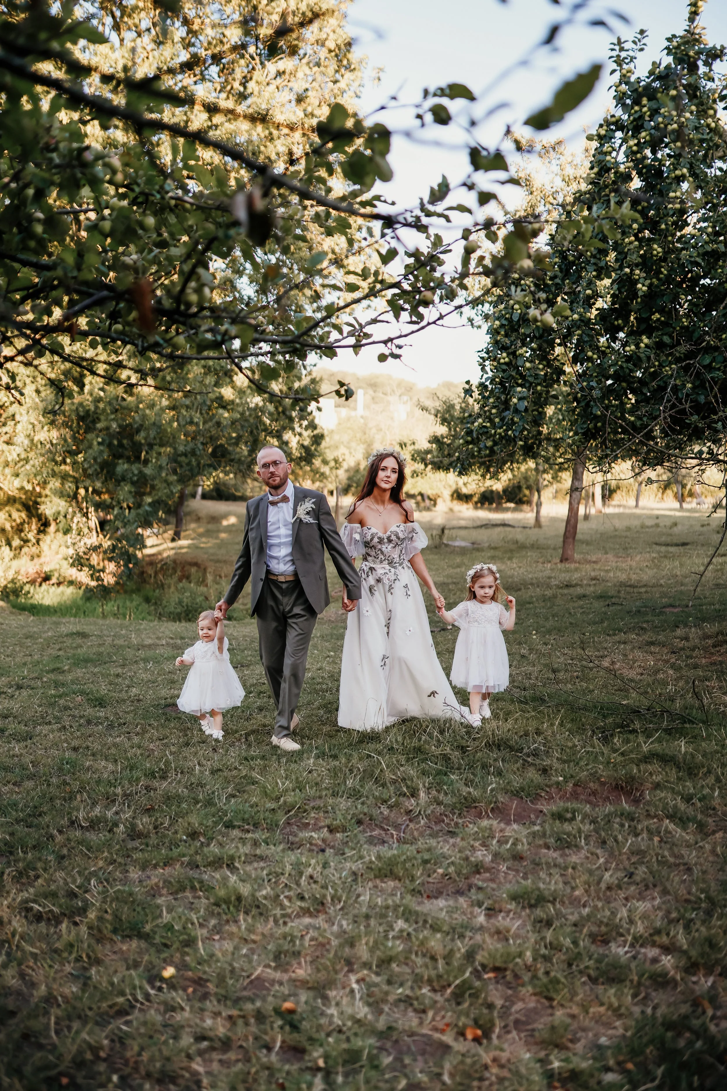A family of four walking hand-in-hand in a park, with the two young girls dressed in white dresses and the adults in formal attire, surrounded by trees and sunlight.