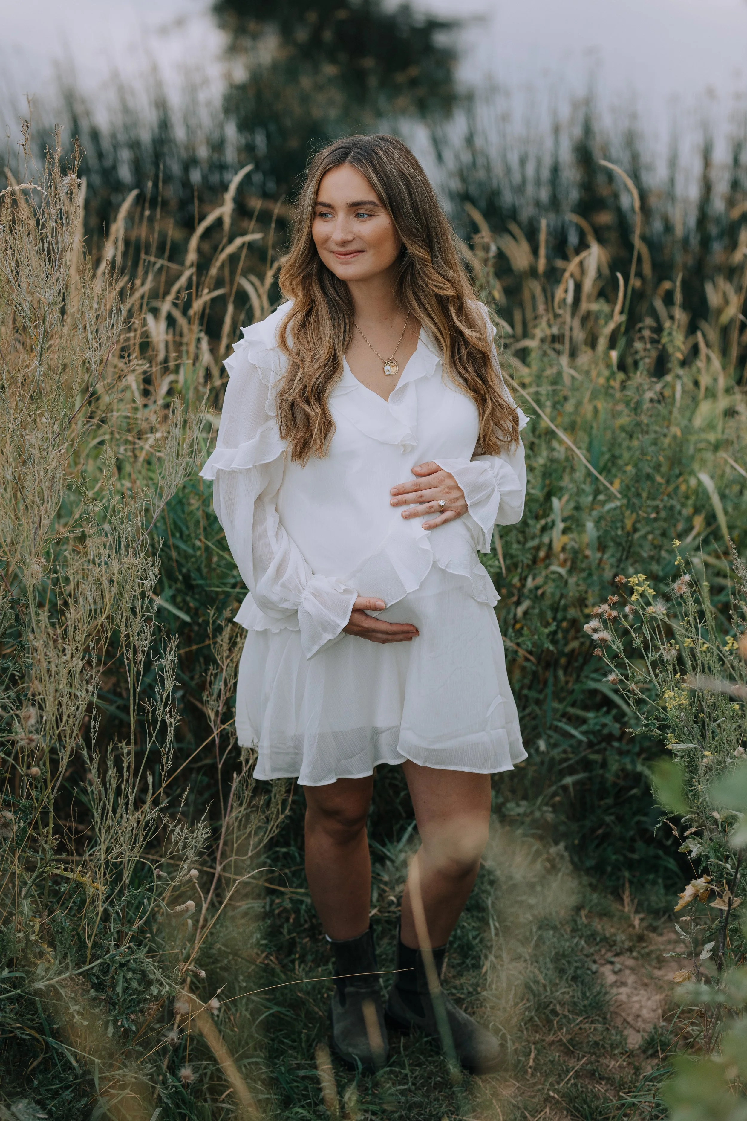 A pregnant woman standing in a field of tall grasses and wildflowers, wearing a white dress, with her hands resting on her belly, and a calm expression on her face.