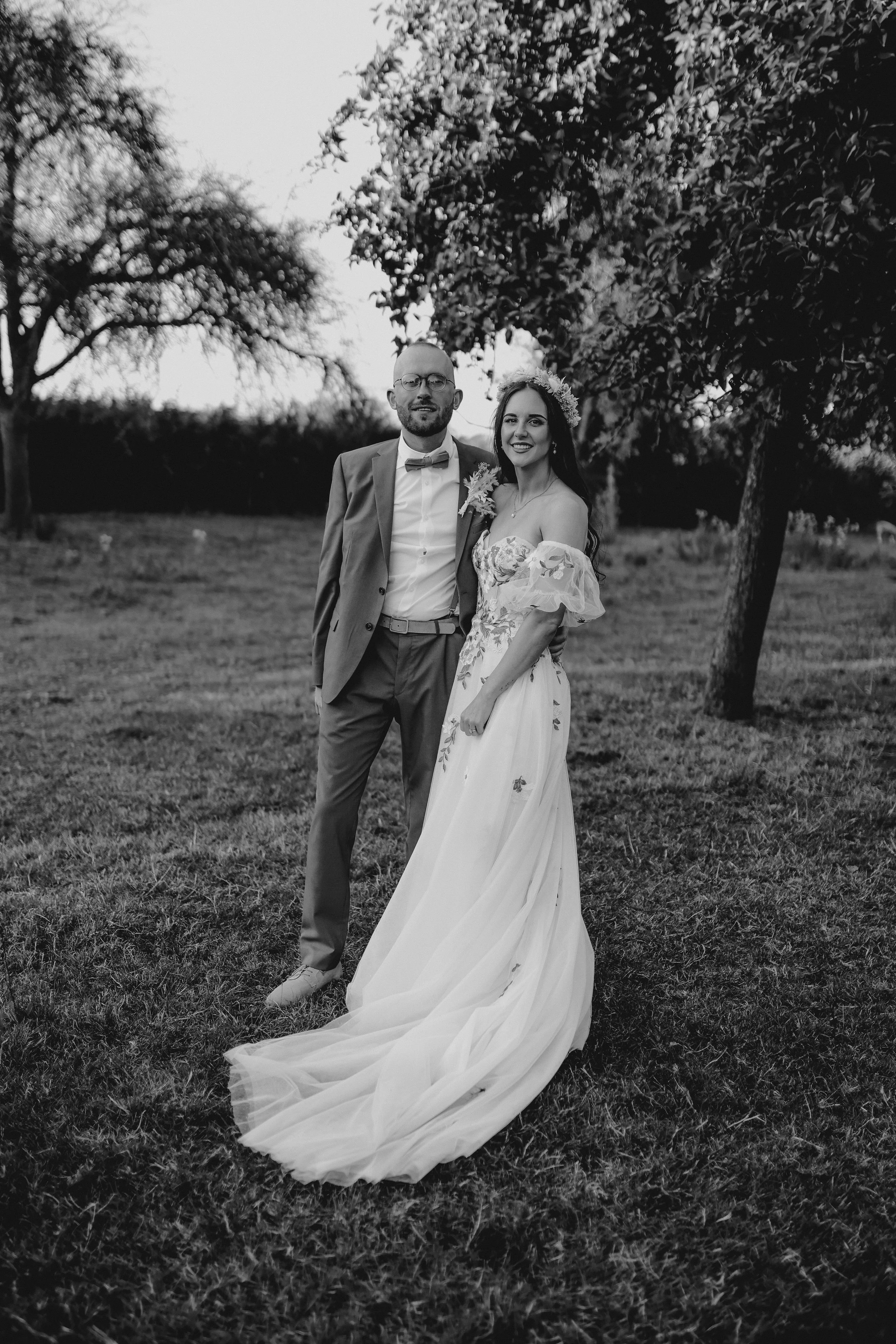 Black and white photo of a bride and groom standing outdoors on grass, with trees in the background. The bride is wearing a wedding dress with off-the-shoulder sleeves and a floral crown, and the groom is in a suit with a bow tie. They are smiling at