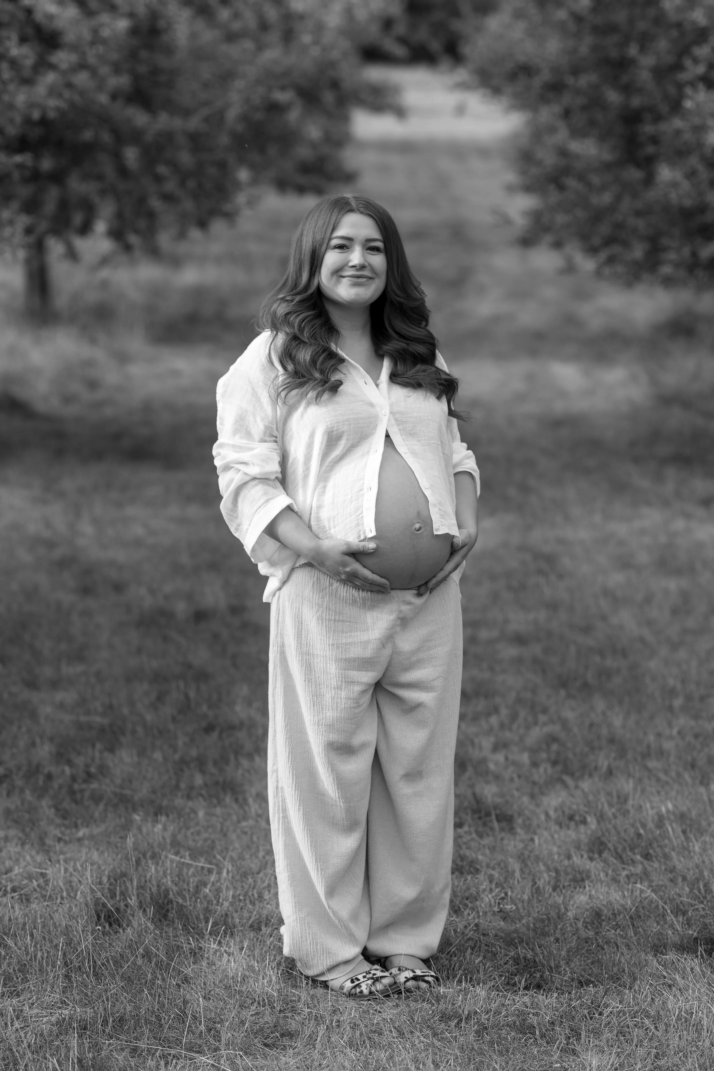 A pregnant woman with long wavy hair standing outdoors in a grassy area, wearing a light-colored open shirt, loose pants, and sandals, with trees in the background