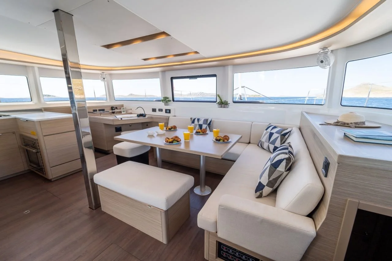 Innenraum einer Yacht mit Esstisch, Stühlen, Sofa, Küchenbereich und Panoramafenstern mit Blick auf das Wasser.