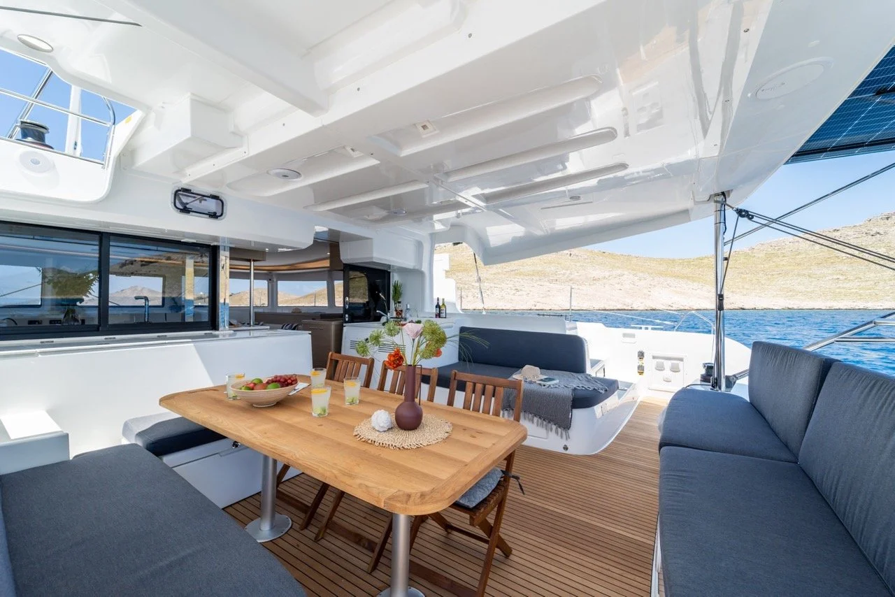Innenansicht einer Yacht mit Essbereich, Meer im Hintergrund, sonniger Tag, moderne Ausstattung, Tisch mit Blumen und Getränken.