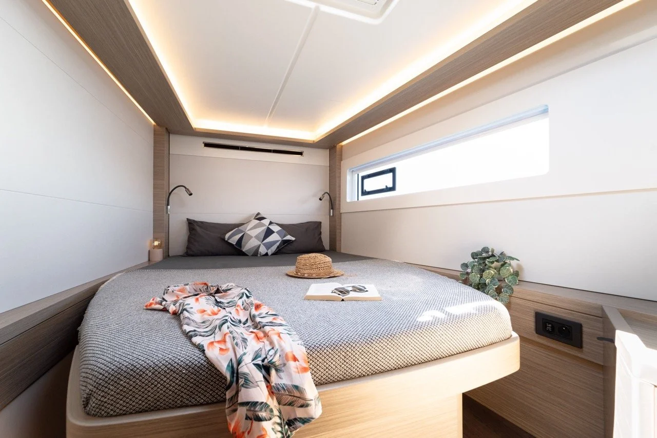 Ein modernes Schlafzimmer mit Doppelbett, grauen Kissen, einer Sonnenhut, einem offenen Buch, einer Pflanzen und einer langen rechteckigen Fenster, mit eingebauter Beleuchtung in der Decke.