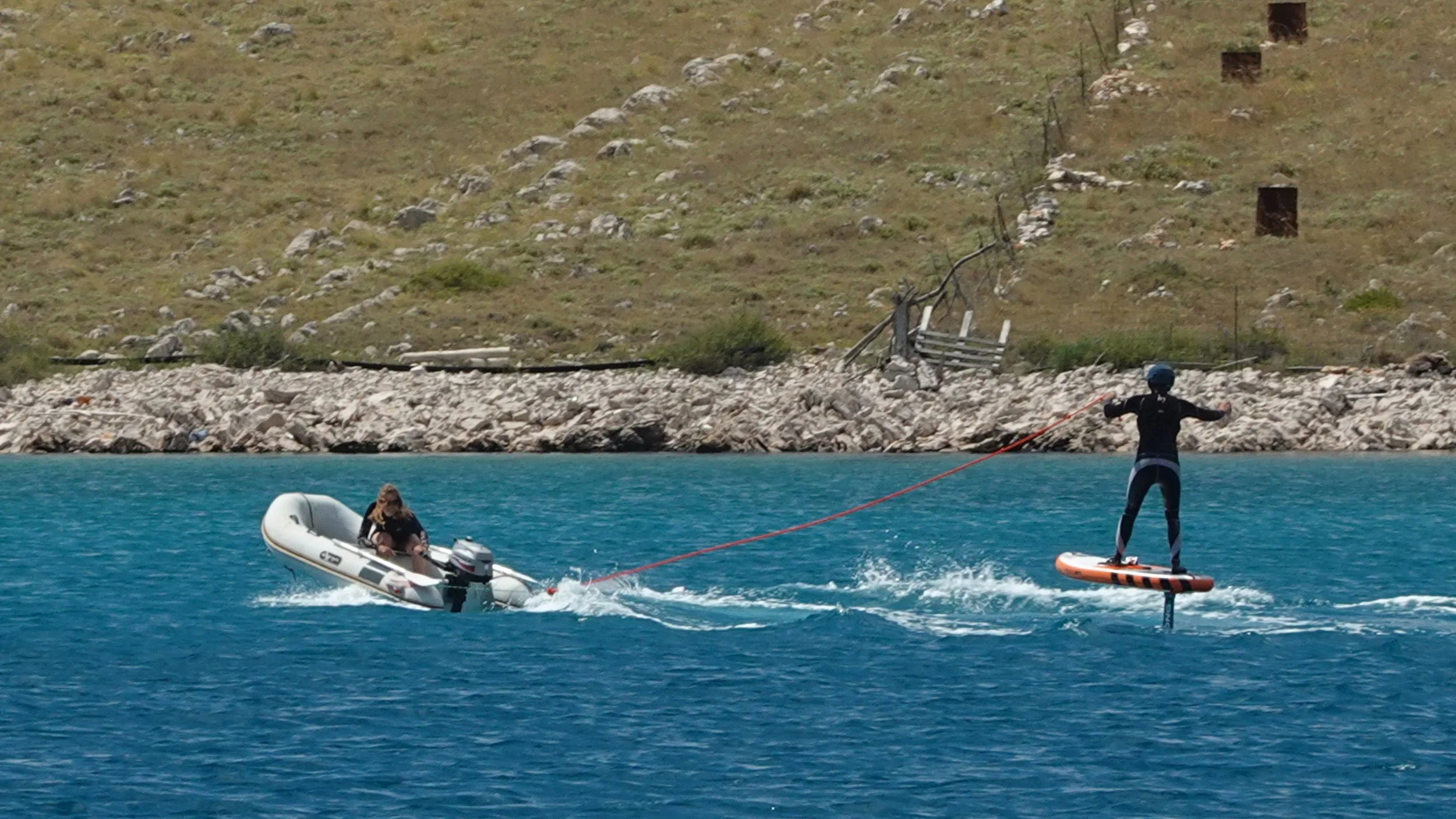 Person auf einem Wasserfahrzeug, das auf Wasserskiern steht, gezogen von einem kleinen Boot im See, mit einer bergigen Landschaft im Hintergrund.