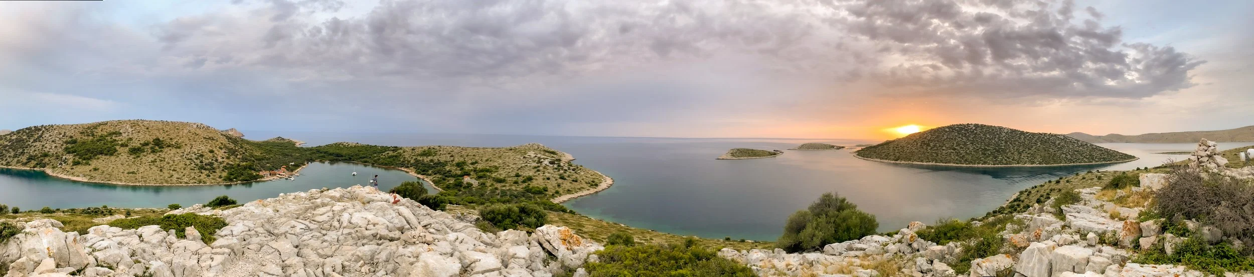 Panoramablick auf eine Lagune mit mehreren Inseln bei Sonnenuntergang, leichter bewölkter Himmel, felsiger Aussichtspunkt im Vordergrund.