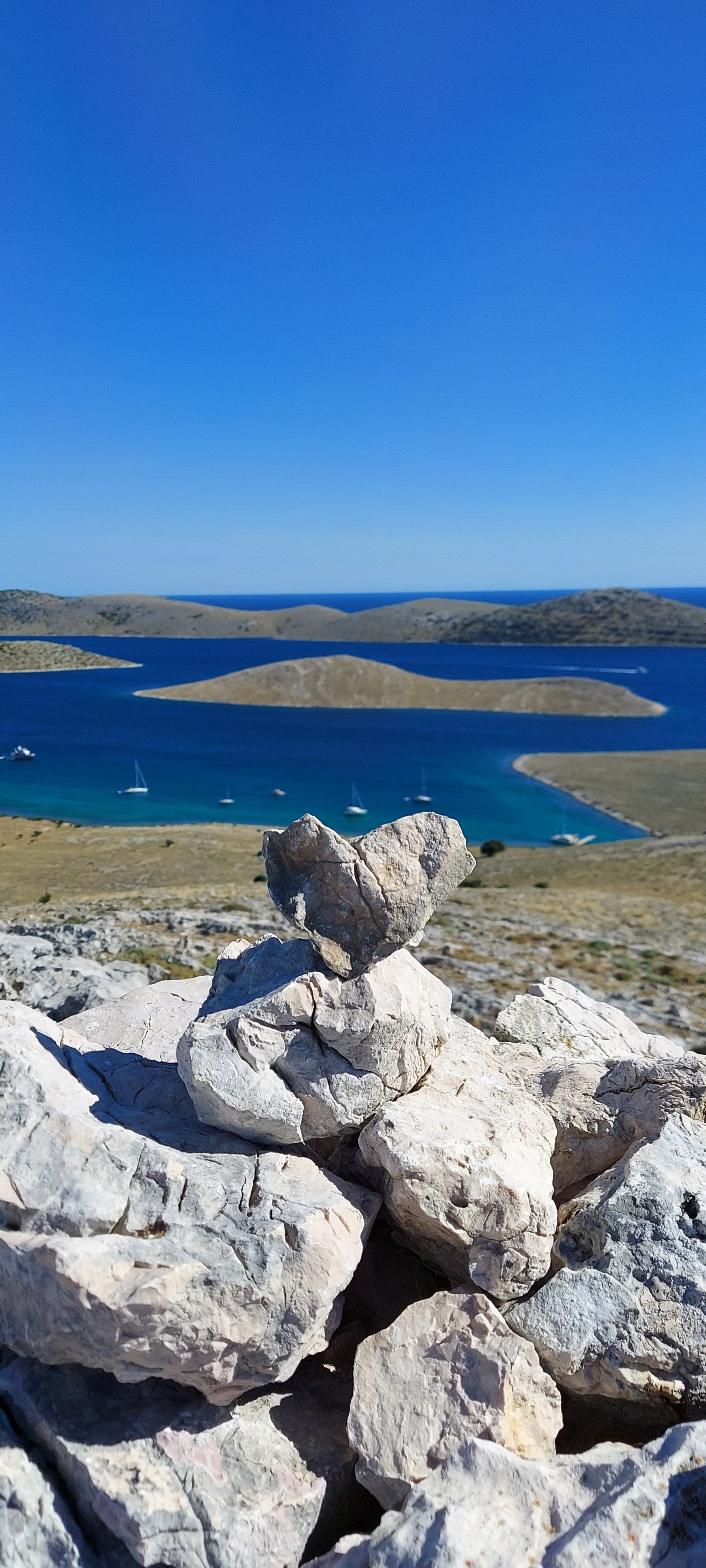 Berggipfel mit Steinen im Vordergrund, Blick auf eine Insel mit Booten im blauen Wasser, klarer Himmel.