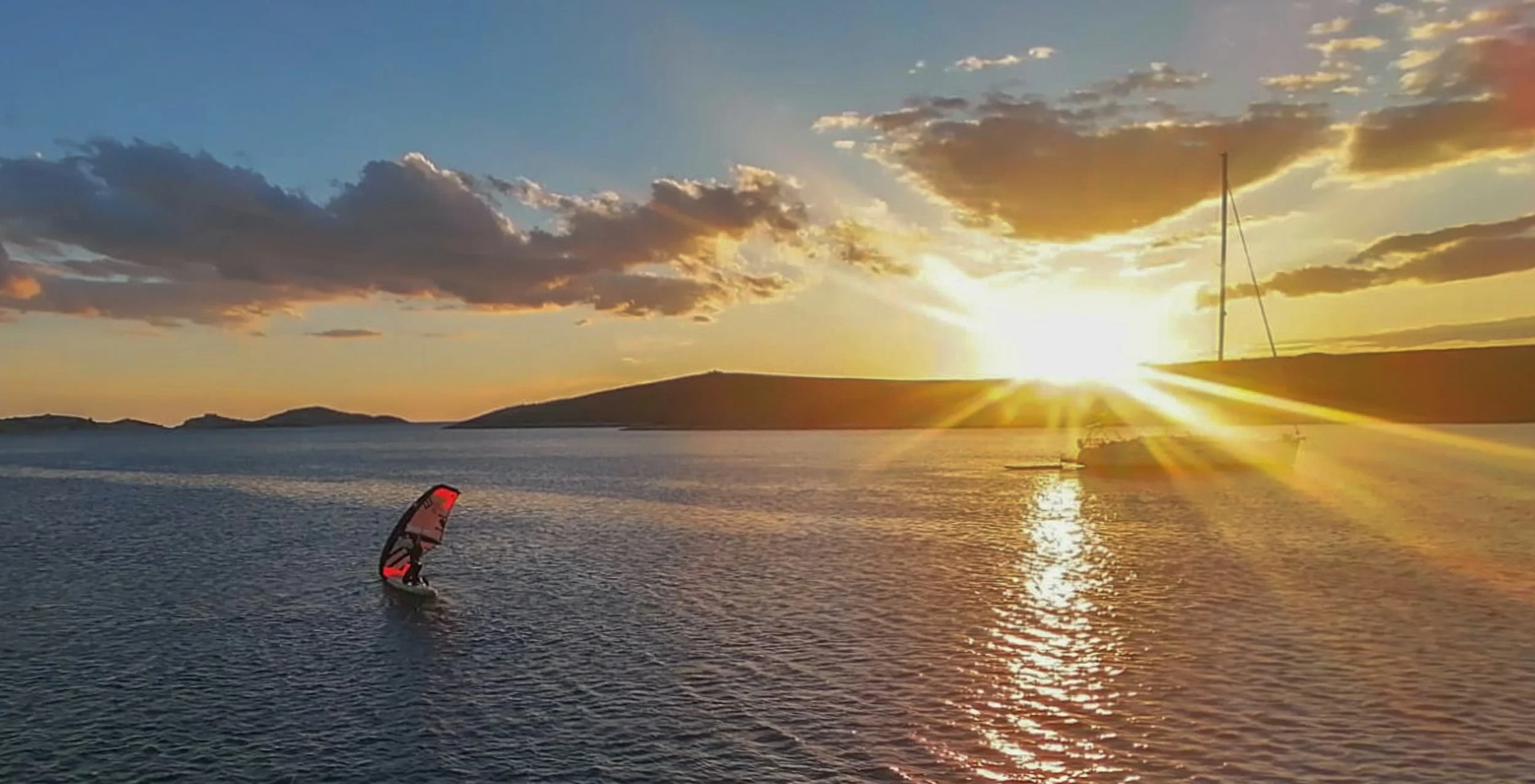 Sonnenuntergang über einem Wassersee, mit einem Windsurfer und einem Segelboot im Vordergrund.
