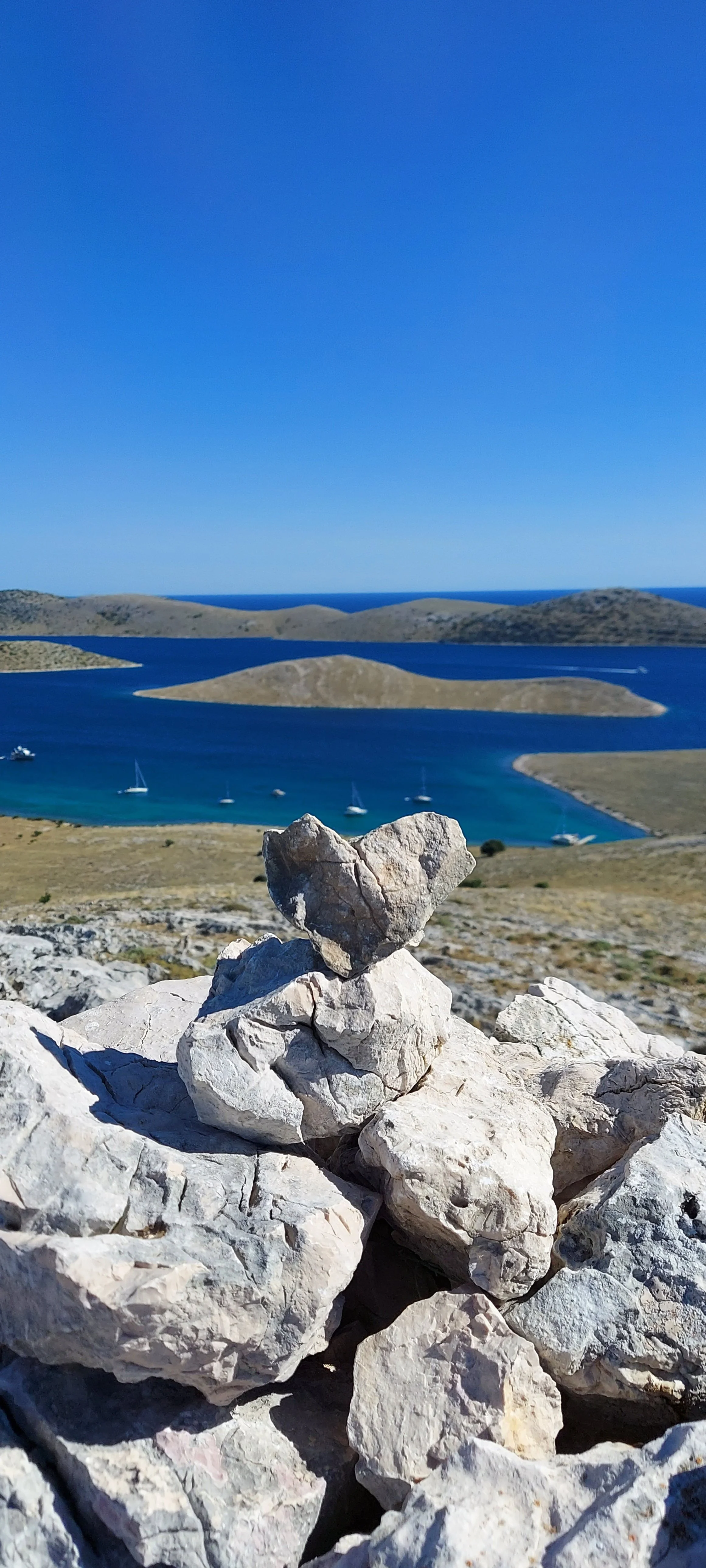 Felsen auf einer Anhöhe mit Blick auf einen See mit mehreren Inseln und Segelbooten, blauer Himmel.