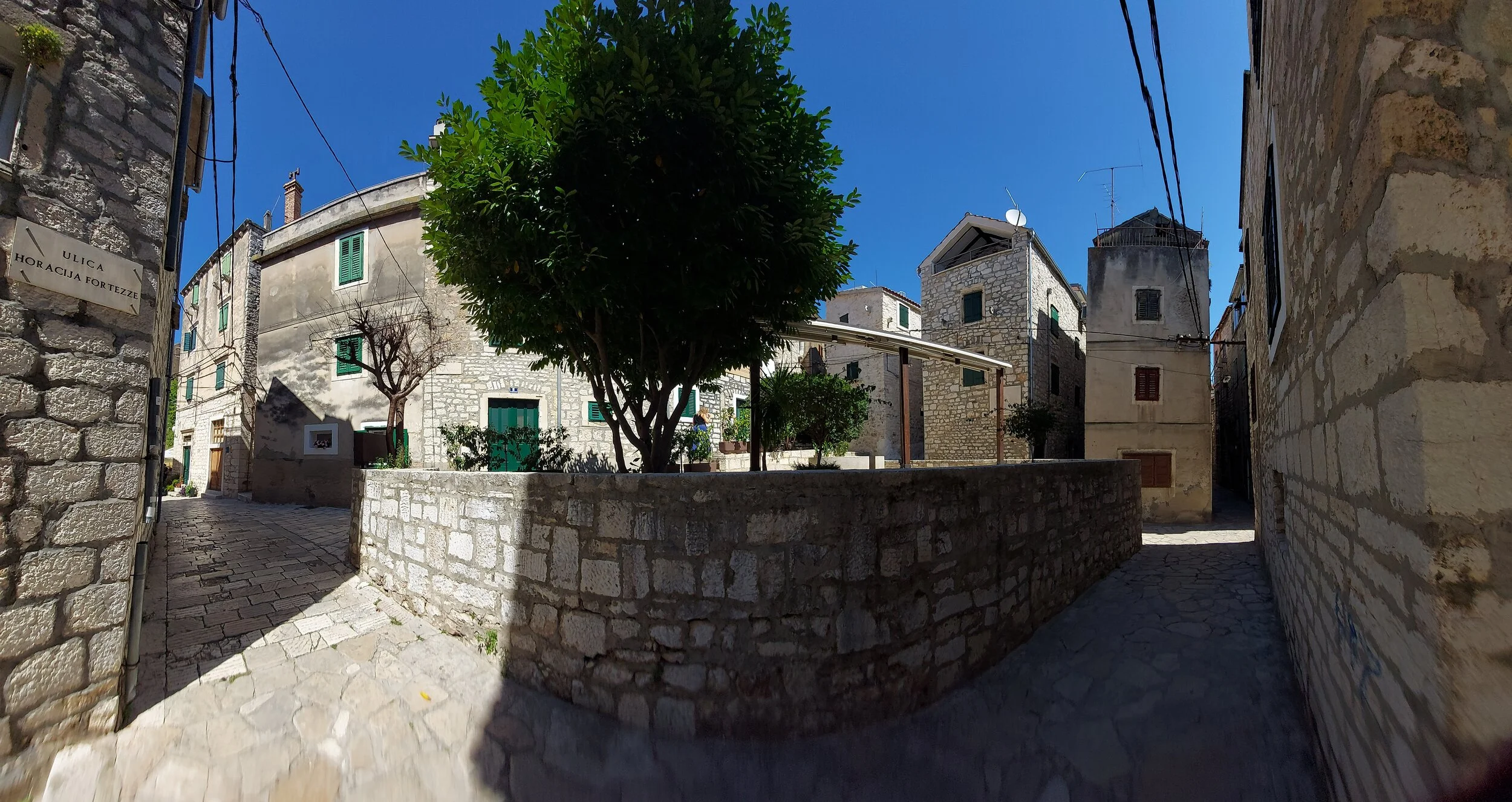 Eine enge Gasse in einem alten Dorf mit Steinmauern, einer großen Baum im Zentrum und mehreren Gebäuden mit grünen Türen und Fenstern. Der Himmel ist klar und blau.