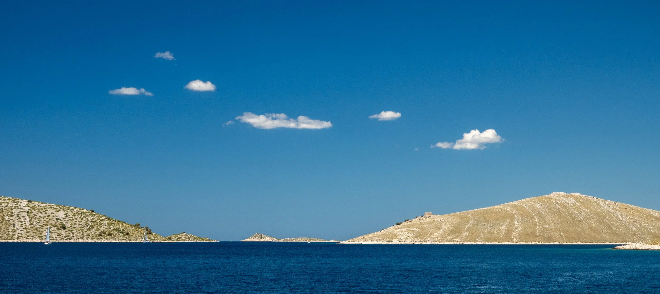 Blick auf eine große Wasserfläche mit einer Insel im Hintergrund, teils bewaldet, und kleinen Segelbooten auf dem Wasser, gegen einen blauen Himmel mit wenigen Wolken.