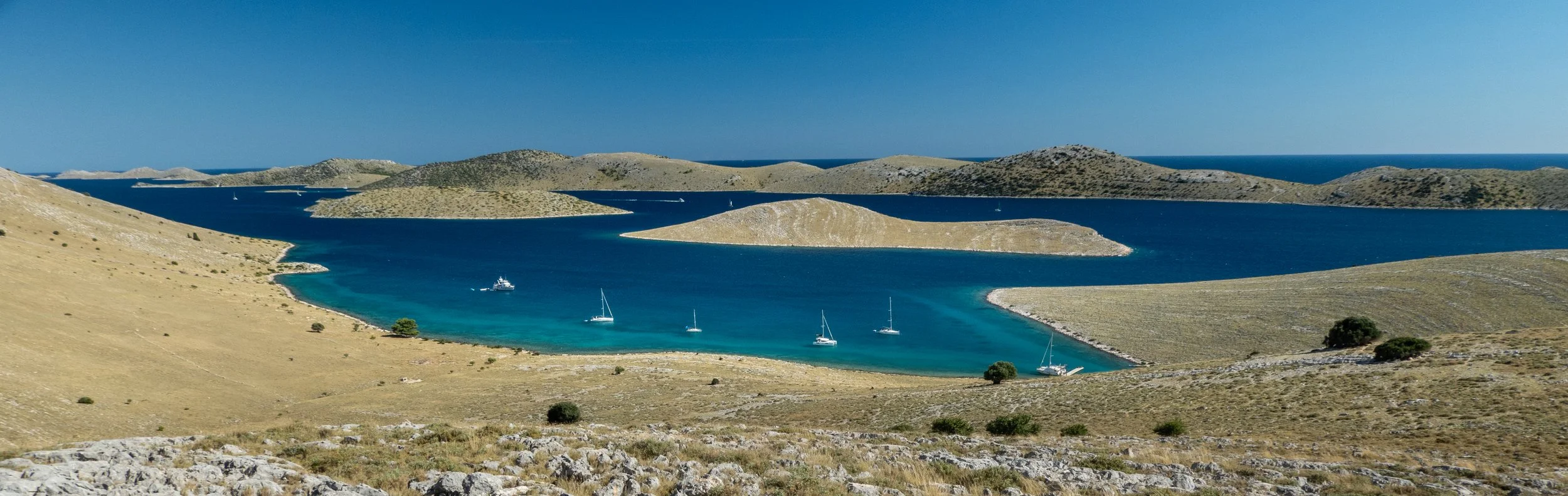 Blick auf einen ruhigen See mit mehreren Segelbooten, umgeben von hügeligem, trockenem Landschaftsgebiet unter blauem Himmel.