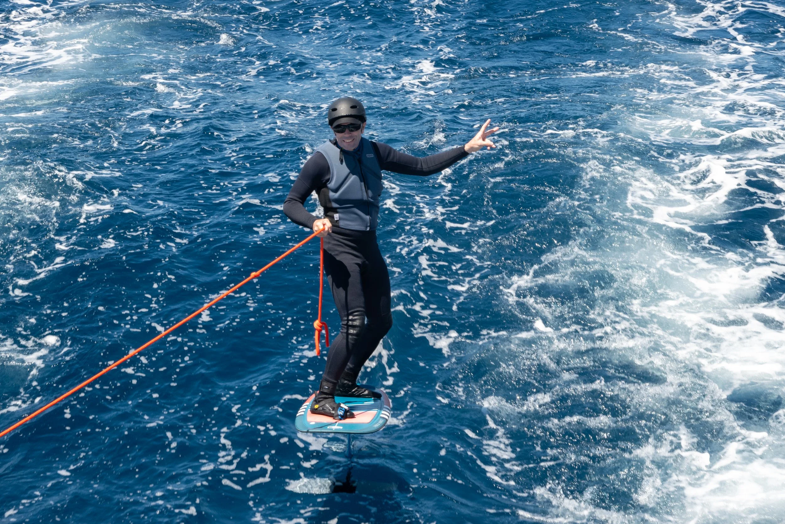 Eine Person beim Wakesurfen auf dem Meer, trägt einen Helm und eine Schwimmweste, zeigt eine Peace-Geste mit der Hand.
