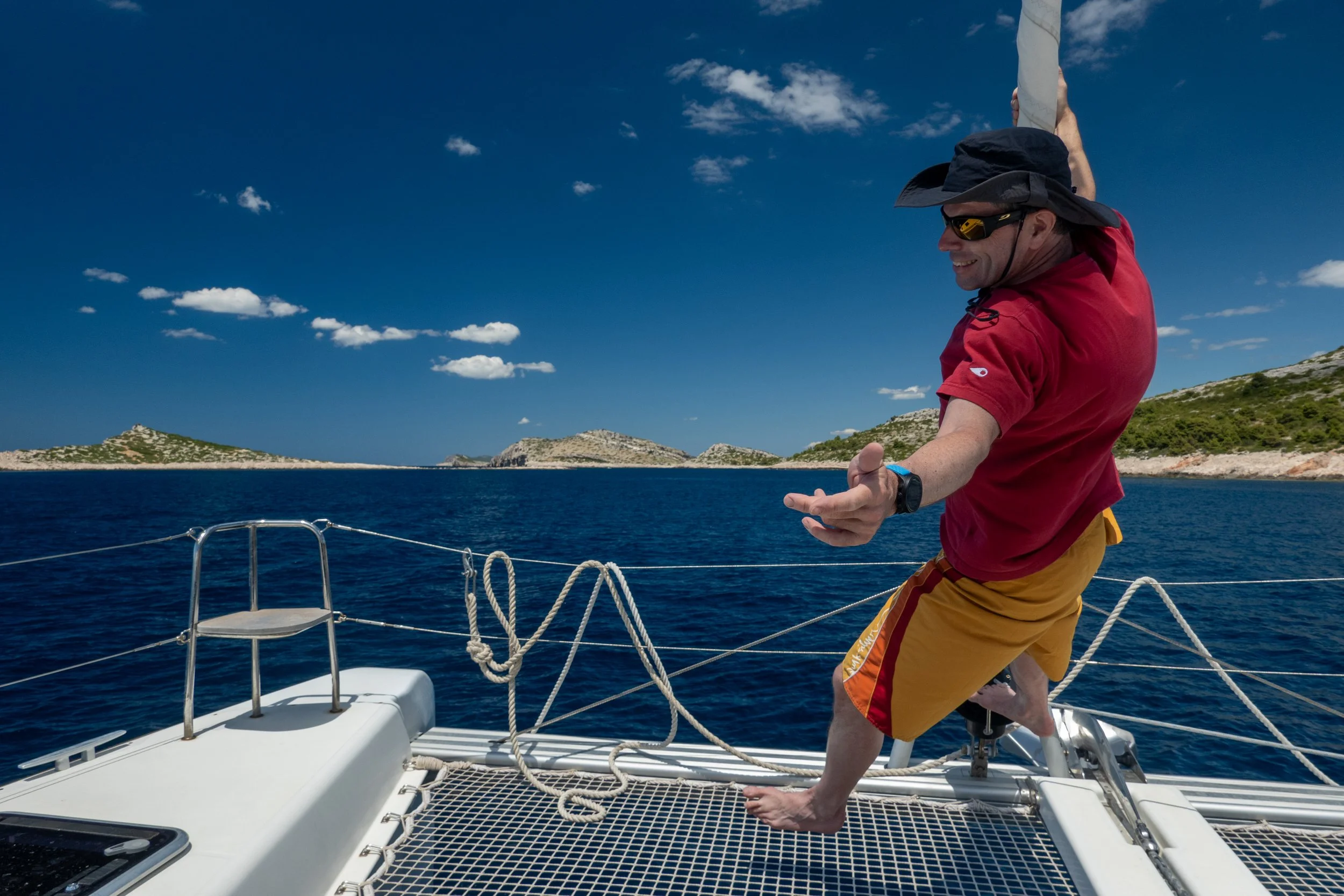 Ein Mann in Sonnenbrille, rotem T-Shirt und gelben Shorts balanciert auf dem vorderen Trampolin eines Katamaran Segelboots auf offener See, mit kleinen Inseln im Hintergrund und blauem Himmel.