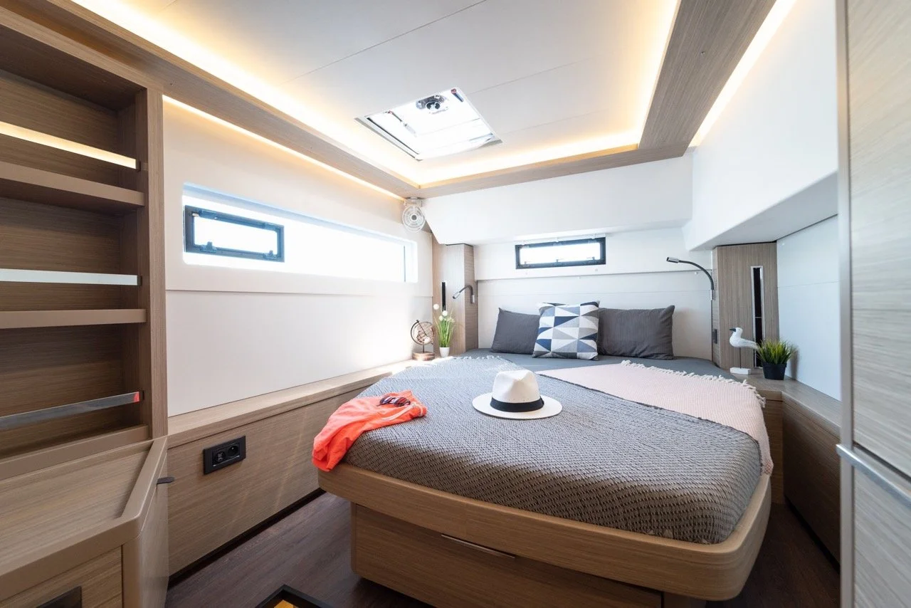 Ein modern eingerichtetes Schlafzimmer mit einem großen Bett, dekorativen Kissen und einer weißen Hut auf der Bettdecke. Es gibt zwei kleine Fenster, eine minimalistische Beleuchtung an der Decke und ein dekoratives Pflanzenelement.