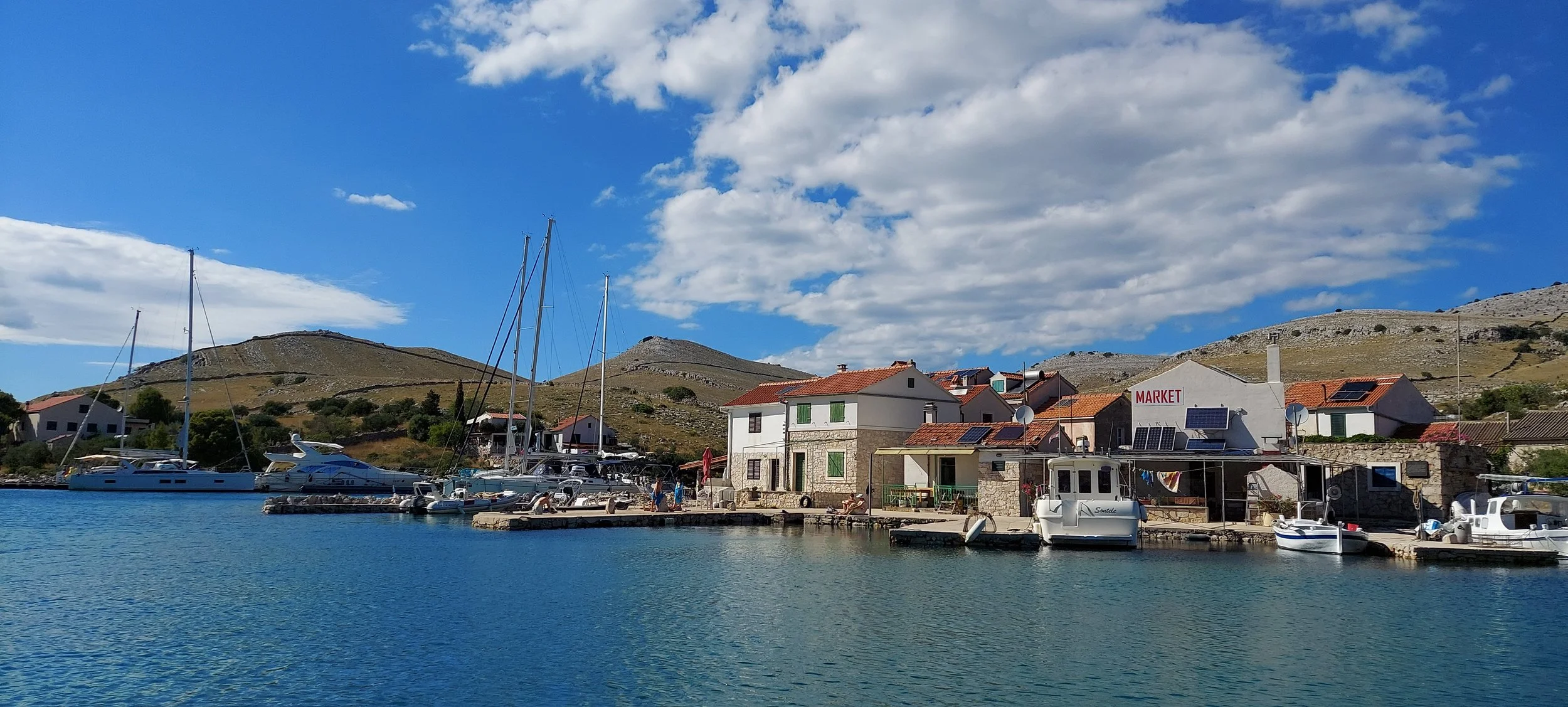 Blick auf eine kleine Hafenstadt mit Booten im Wasser, umgeben von Häusern mit Solarpanelen, auf Hügeln im Hintergrund, bei sonnigem Himmel mit einigen Wolken.