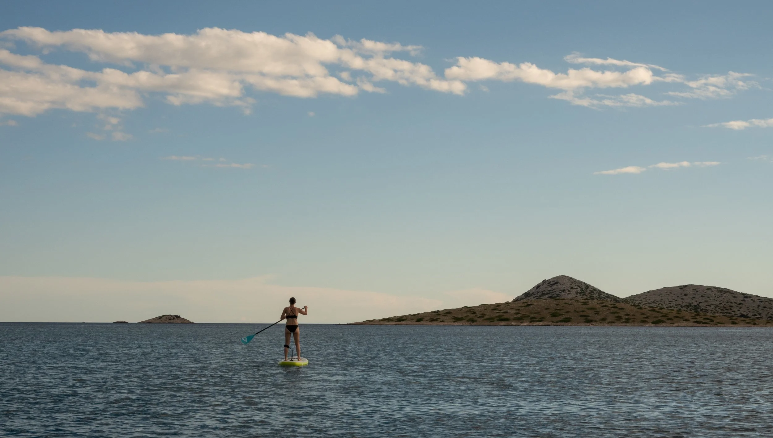 Person auf einem Stand-Up-Paddle-Board in einem ruhigen, abgelegenen Gewässer mit Inseln im Hintergrund bei sonnigem Himmel.