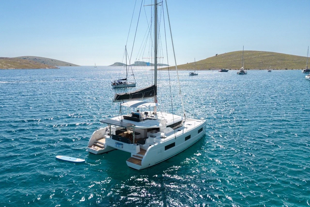 Segelboot auf dem Meer mit mehreren kleineren Booten im Hintergrund und grünen Hügeln an der Küste unter klarem blauen Himmel.