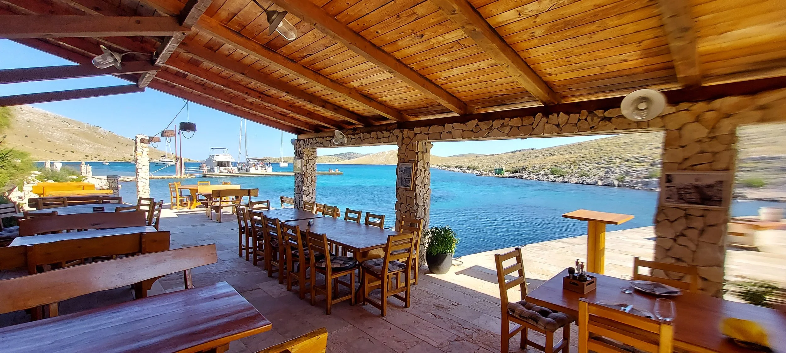 Ein kanarischer Hafen mit Blick auf das Wasser, Boote und Berge im Hintergrund, während im Vordergrund eine offene Restaurantterrasse mit Holztischen und Stühlen zu sehen ist.