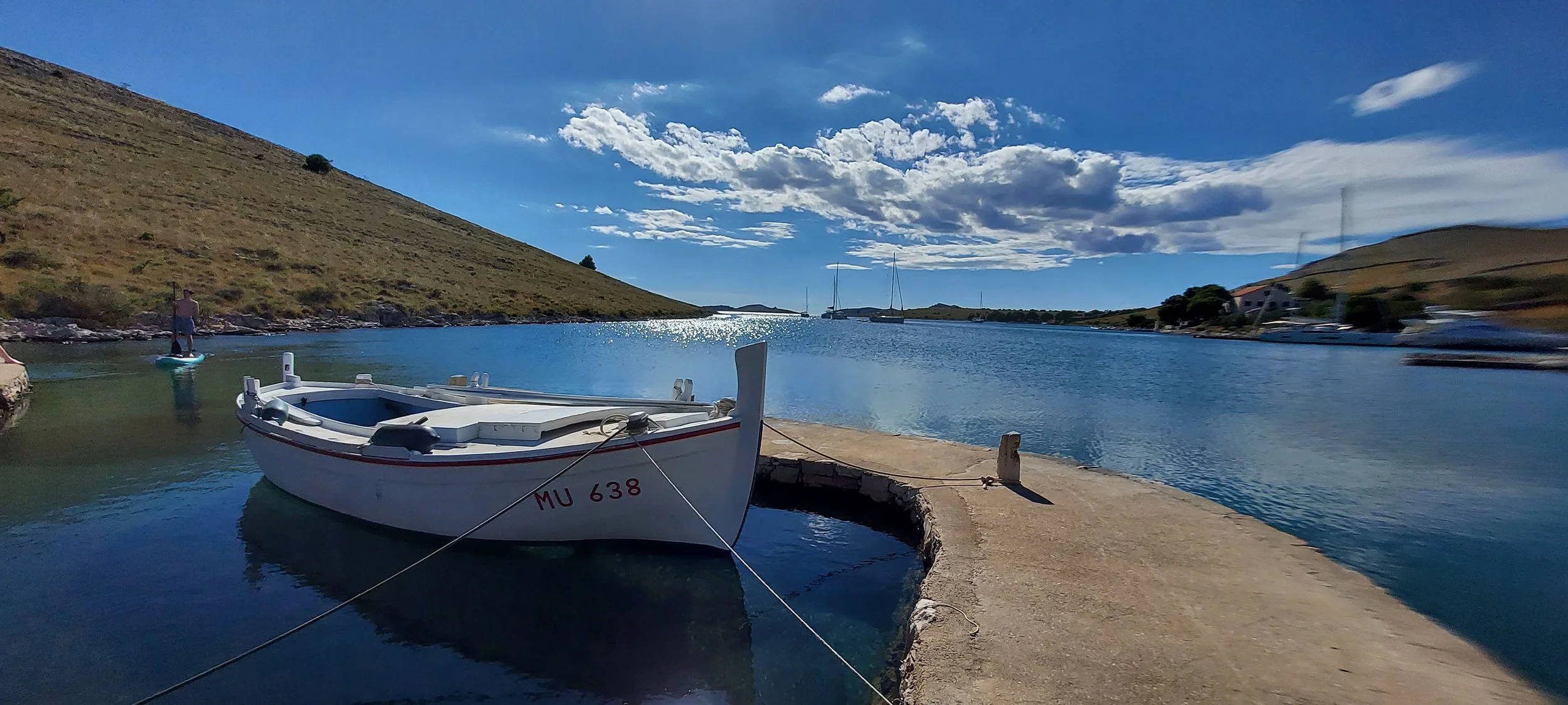 Ein weißes Boot mit roter Markierung am Rumpf liegt am Kai in einem ruhigen Hafen, umgeben von Wasser und sanften Hügeln.
