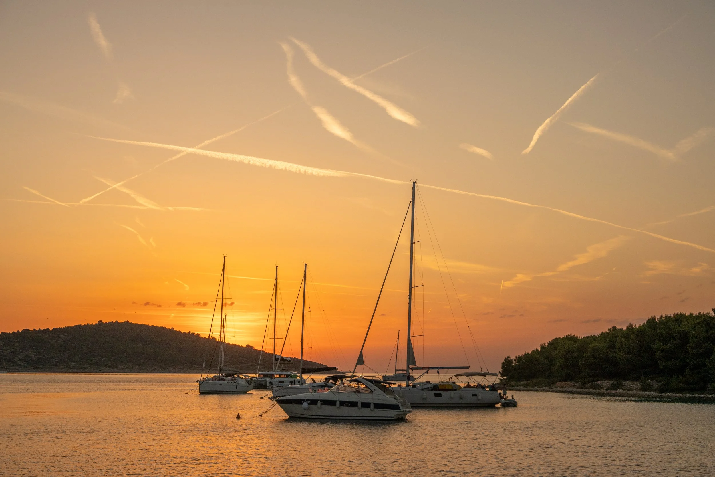 Segelboote auf ruhigem Wasser bei Sonnenuntergang, mit einer bewaldeten Insel im Hintergrund und einem Himmel voller Wolken- und Flugzeug-Wölkchen.