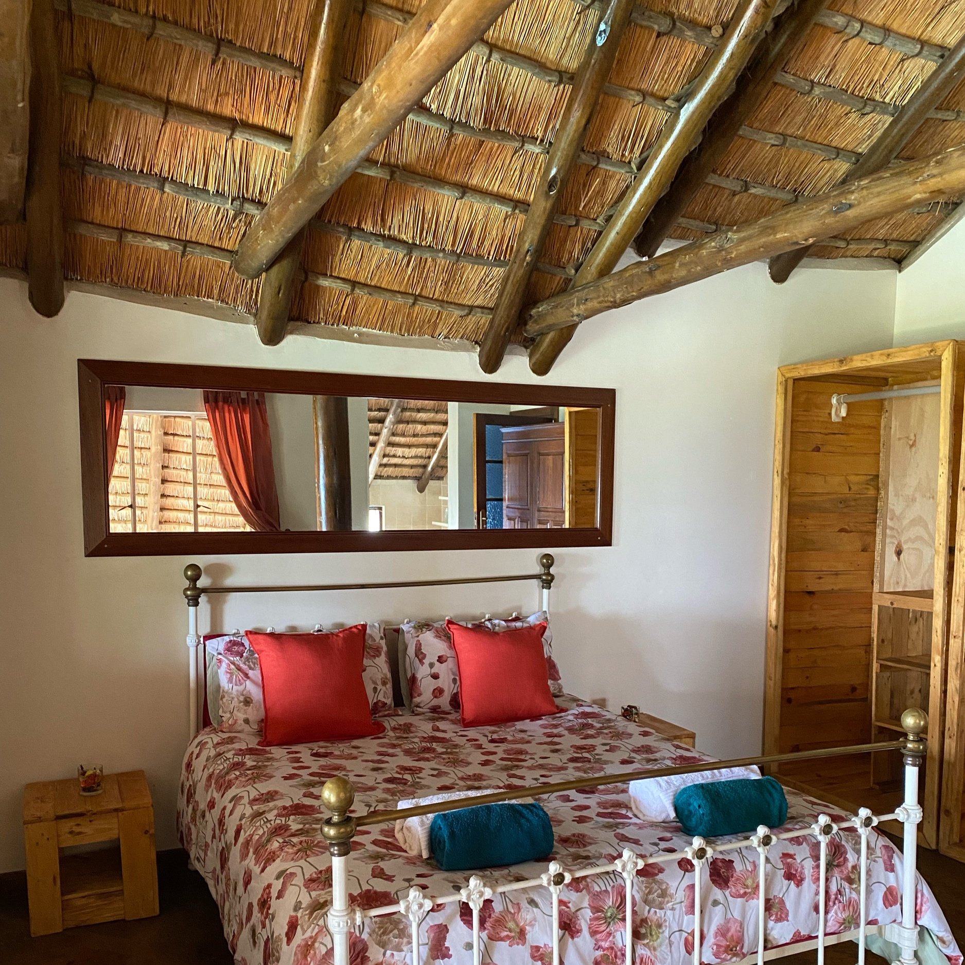 Rustic bedroom with a thatched wooden ceiling, wrought iron bed, floral bedding, red cushions, large mirror, and wooden furnishings.