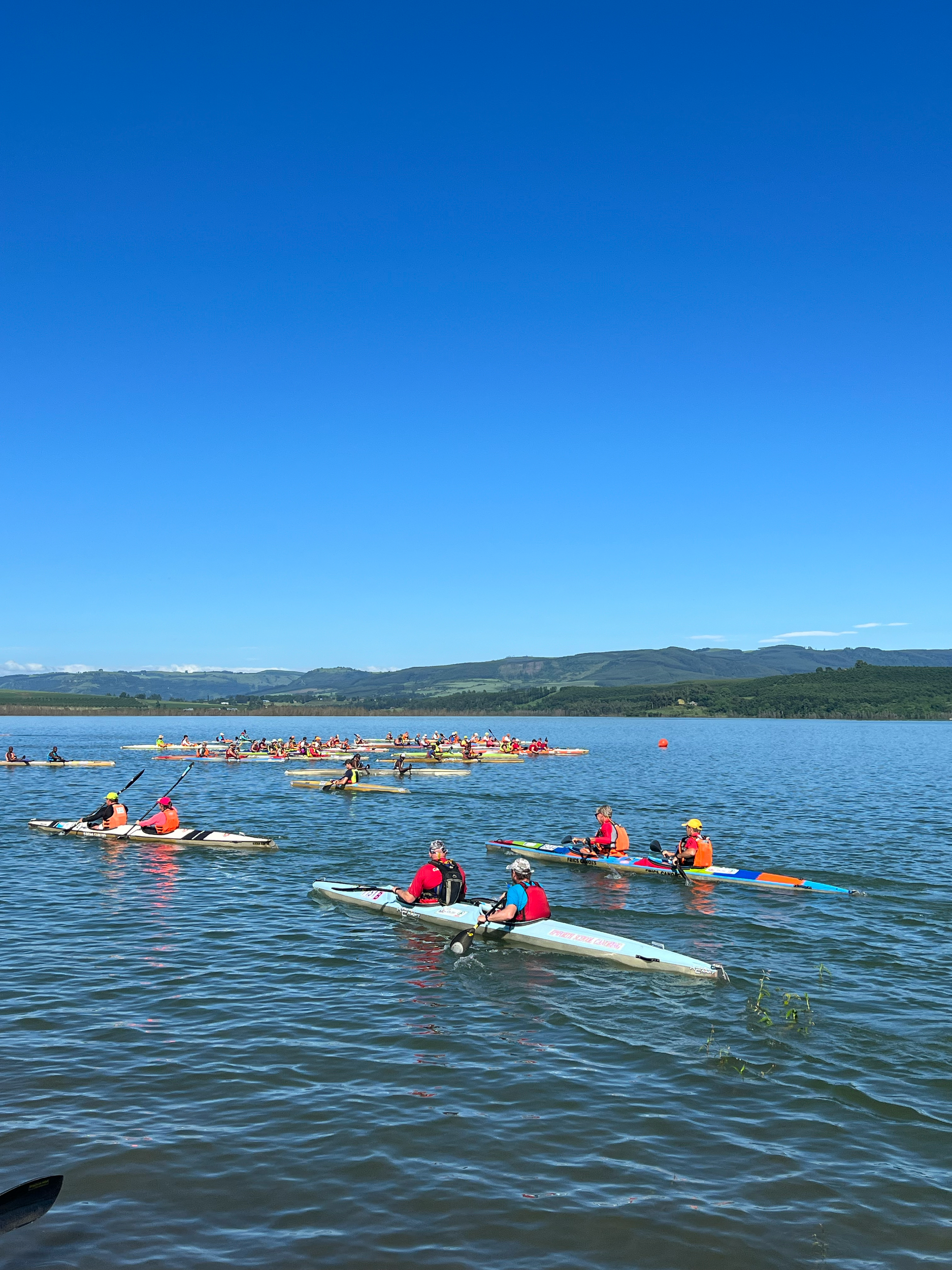 Group of people kayaking on a lake under a clear blue sky, wearing life jackets and helmets, participating in a water event or race.