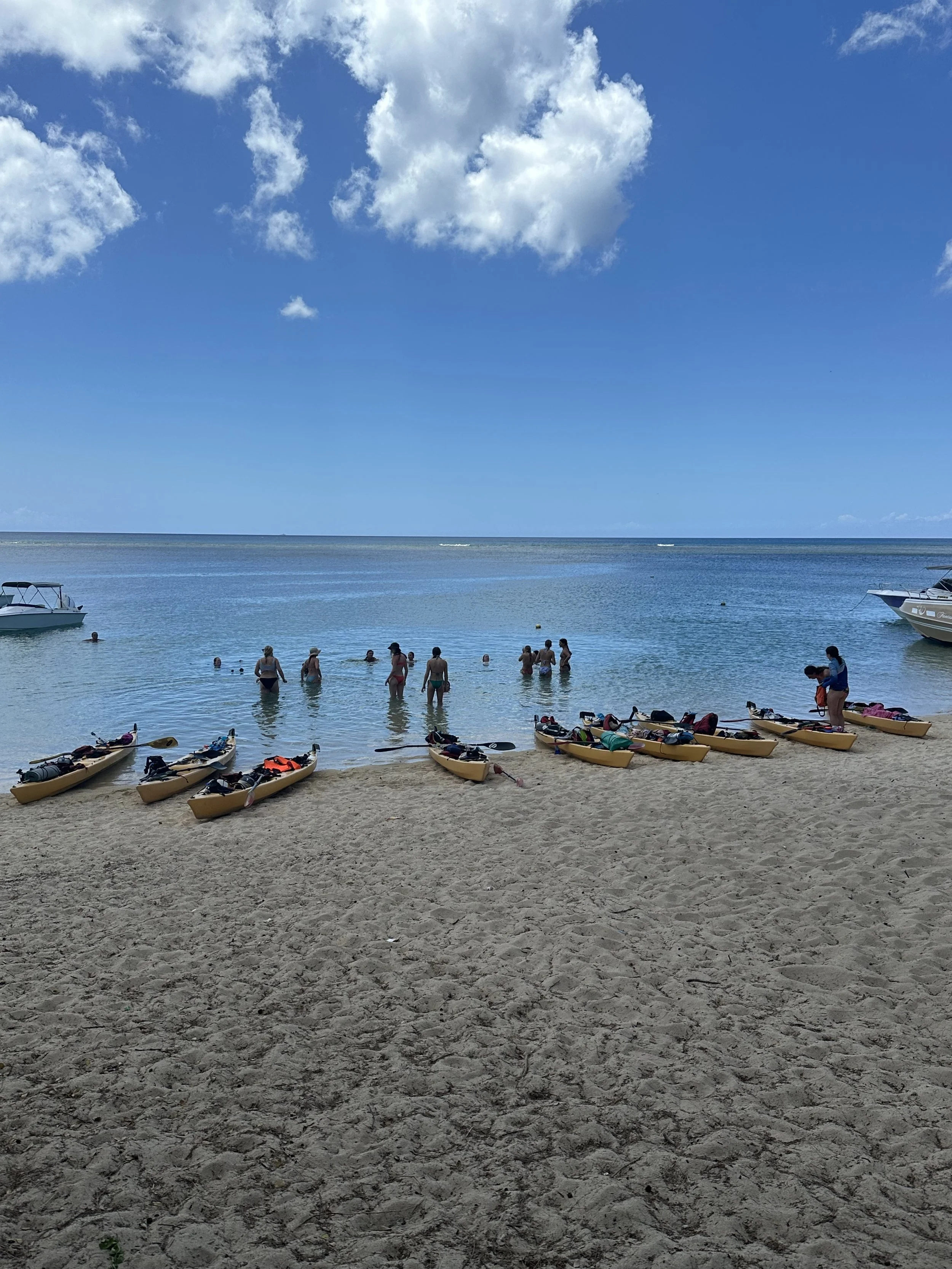 People at the beach with kayaks on the sand and swimmers in the water under a blue sky with clouds.