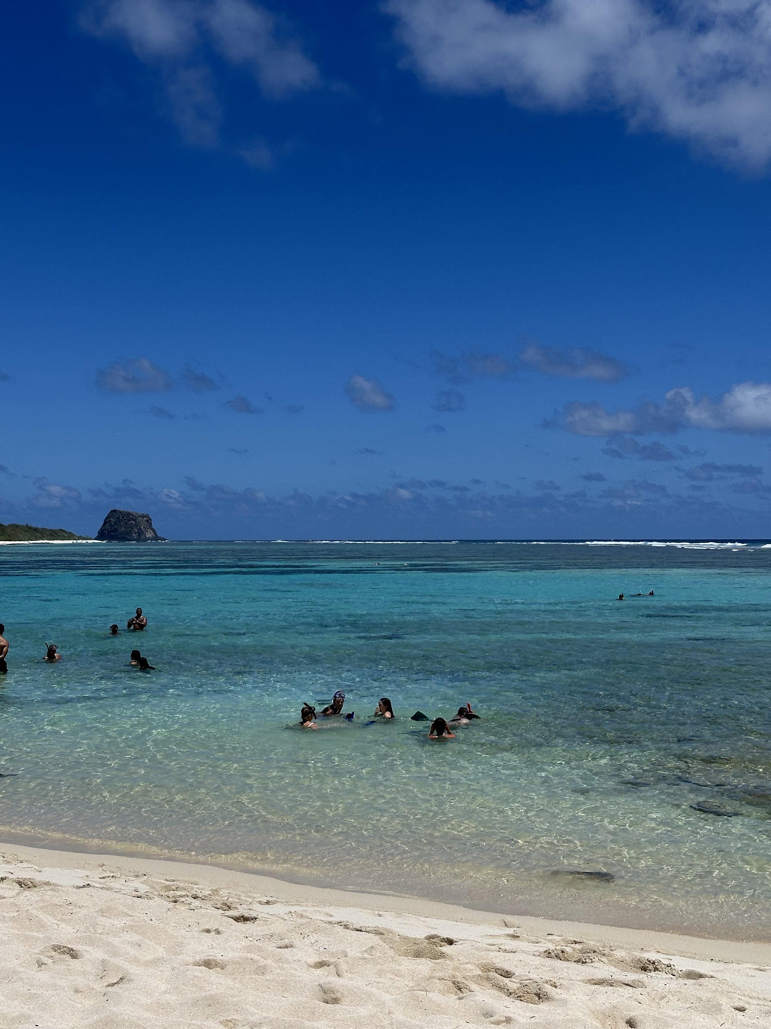 People swimming and relaxing in clear tropical ocean water with sandy beach in foreground and blue sky with some clouds.