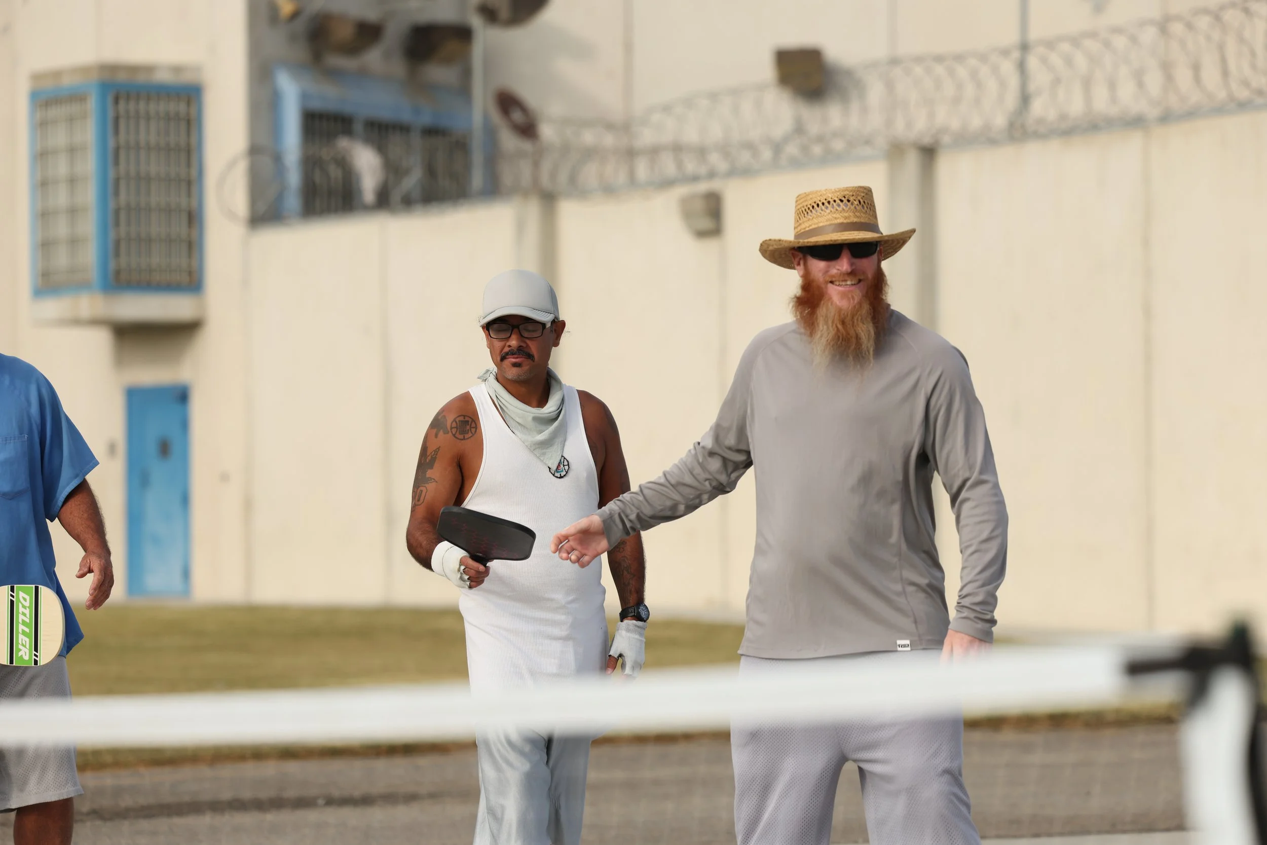 Two men, one with a beard and straw hat, and the other with glasses and a white cap, standing outdoors near a tennis court with a net in the foreground. The bearded man is smiling and gesturing.