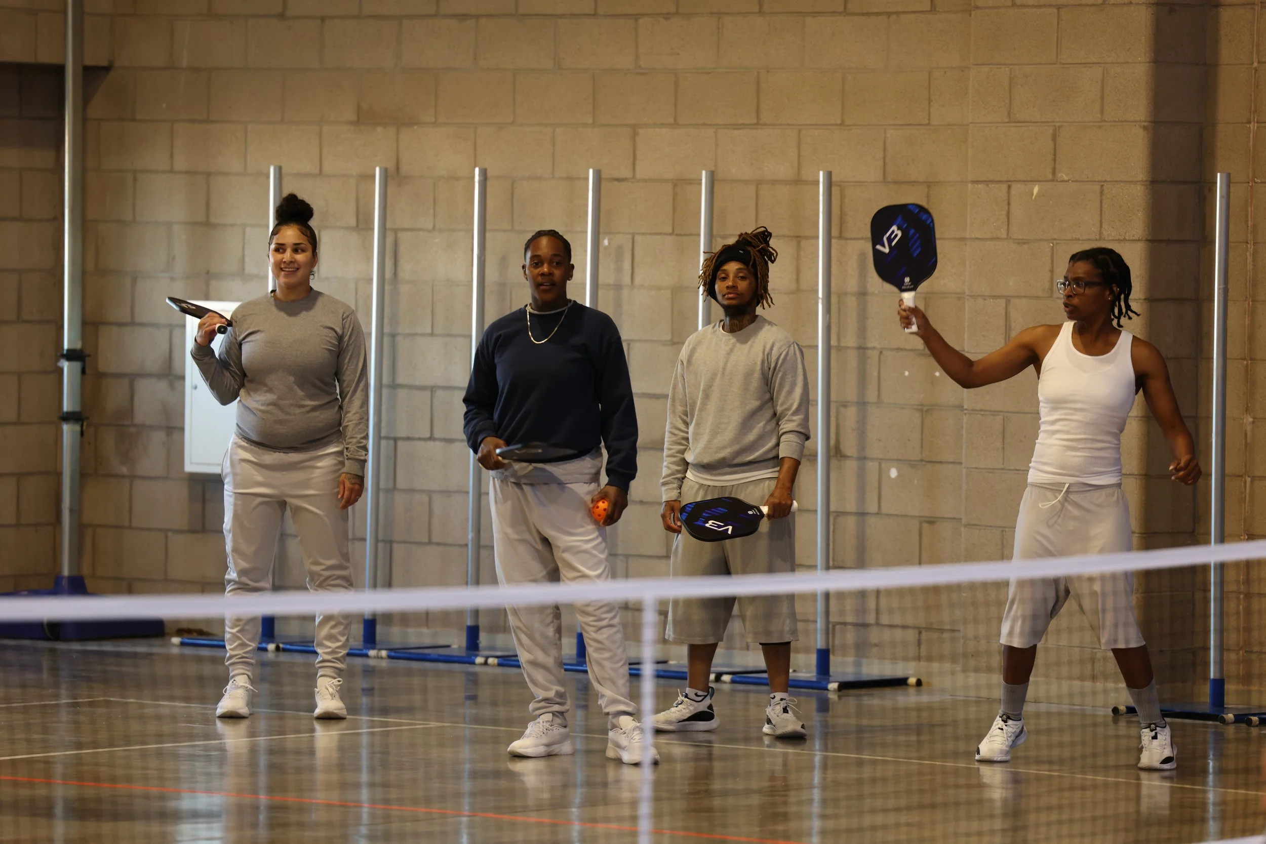 Four female prison inmates stand on an indoor pickleball court with rackets, one is holding a paddle, and a net is in the foreground.