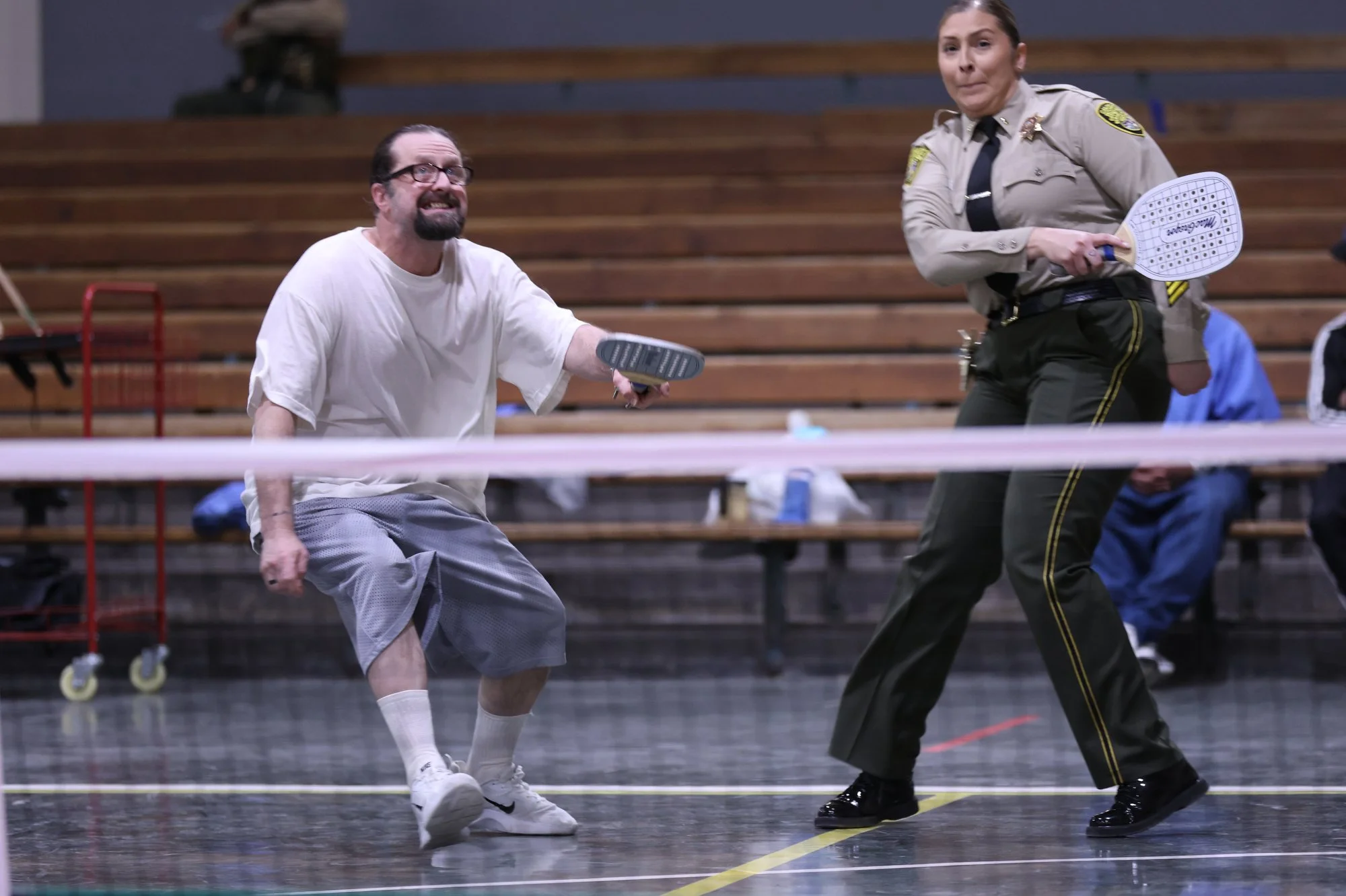 A man in a white t-shirt and shorts has a leg torn and is missing a shoe, playing tennis against a woman in a police uniform at an indoor tennis court with wooden bleachers in the background.