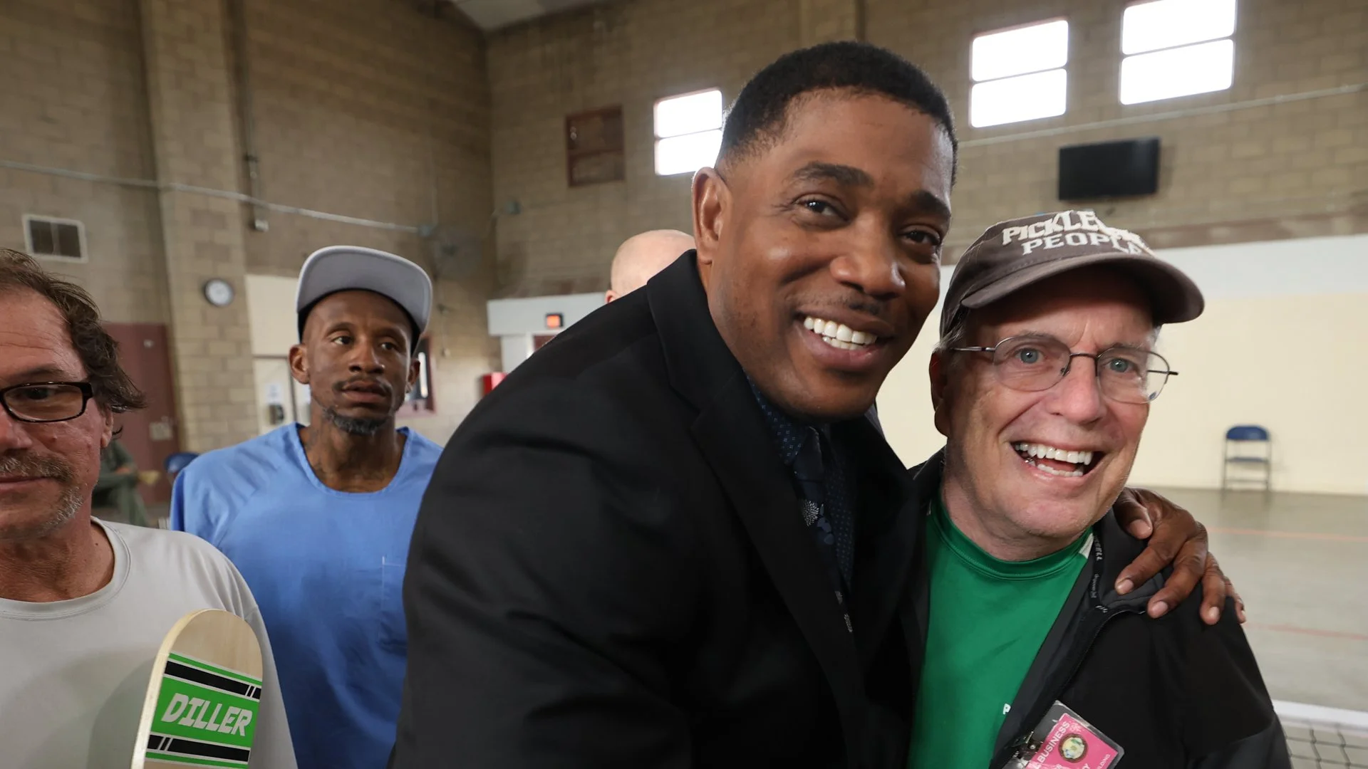 People smiling and posing for a photo indoors, one man has a skateboard, another is wearing a 'Pickle People' hat, and they appear to be at a community event or gathering.