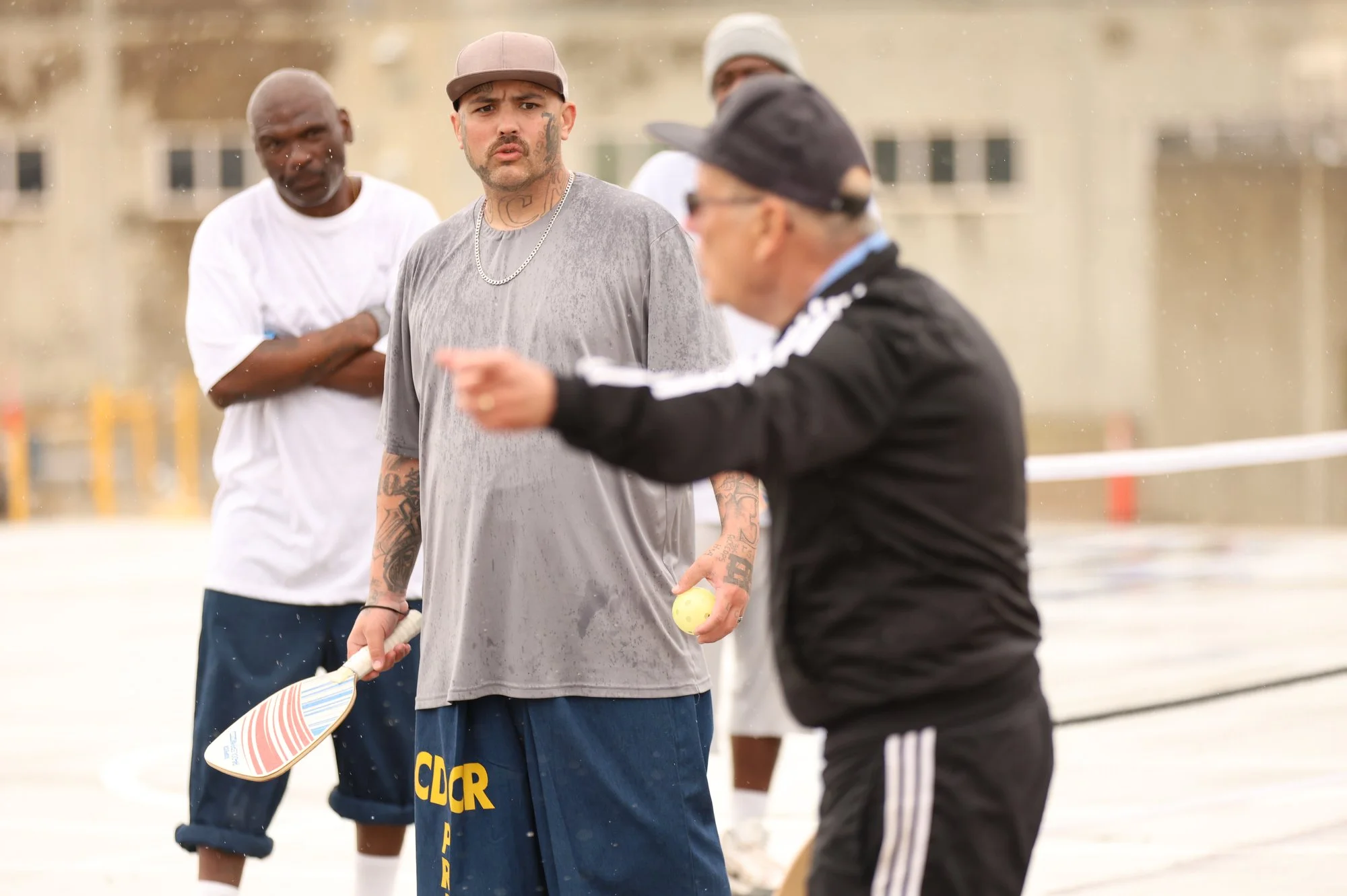 Three men playing pickleball outdoors on a rainy day, one man appears to be instructing or arguing, and another holding a pickleball paddle.