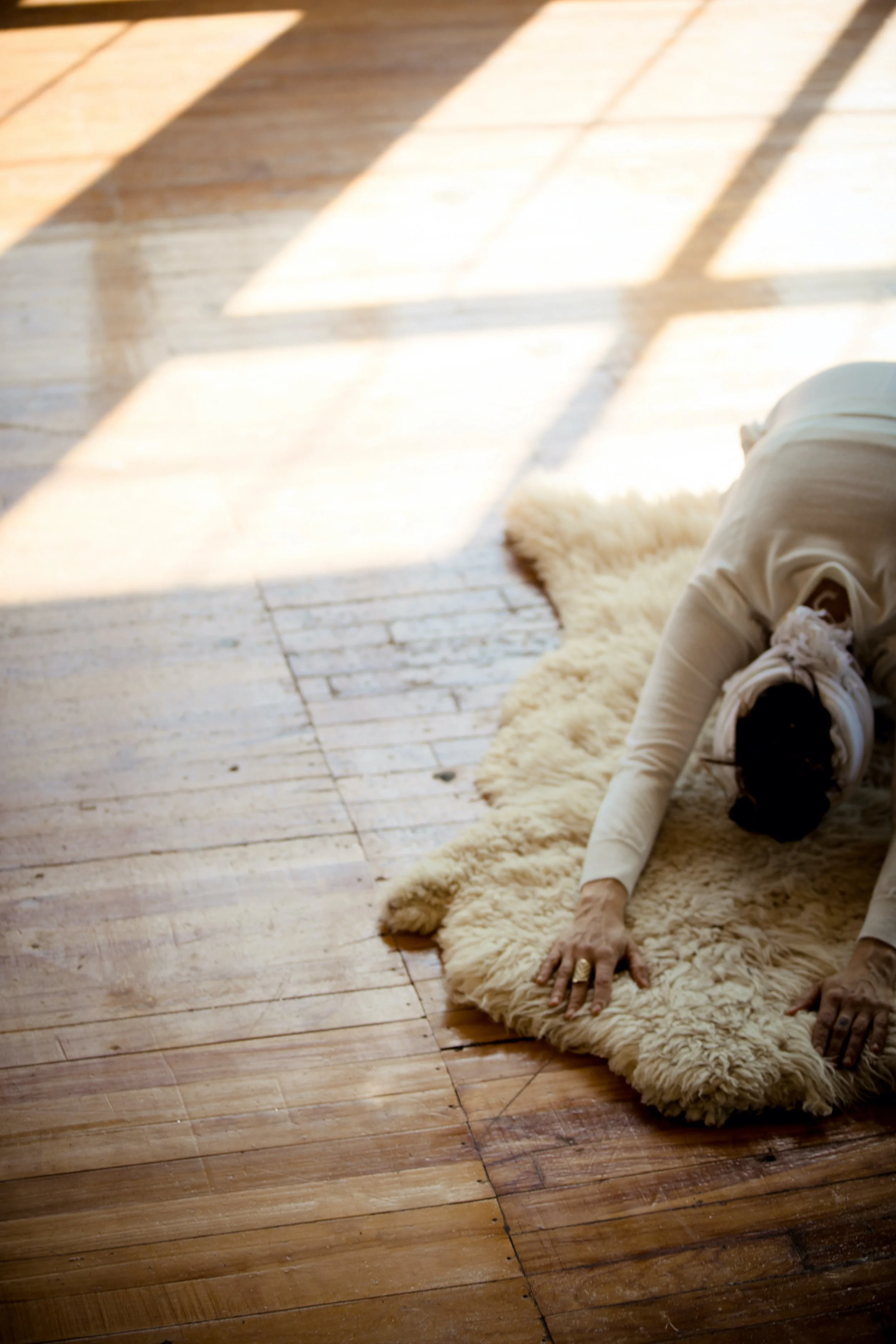 Person in a white hoodie bending over on a fluffy cream-colored rug on a wooden floor with sunlight streaming through a window.