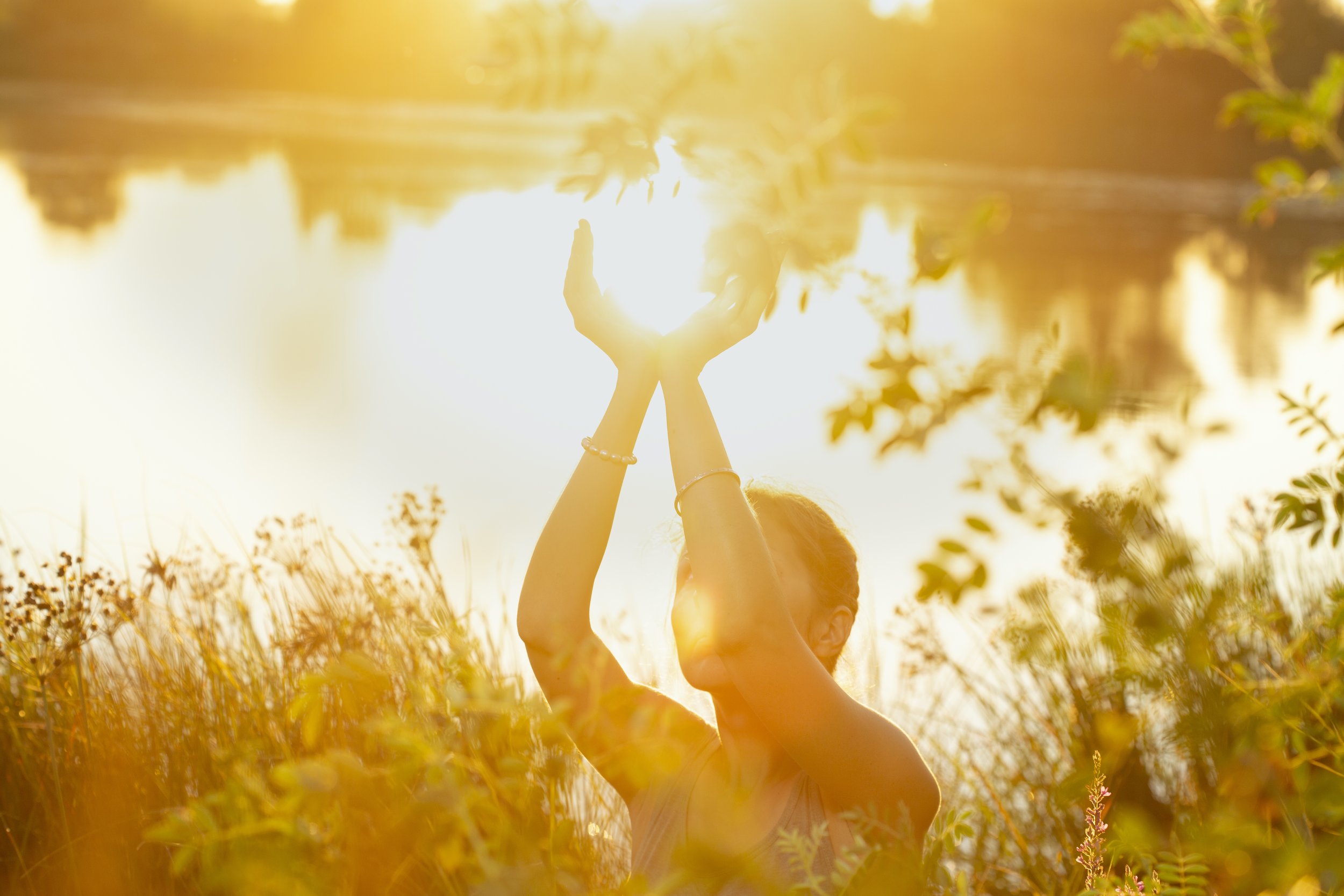 A woman standing outdoors near a lake during sunset, with her hands raised towards the sky, catching the sunlight.