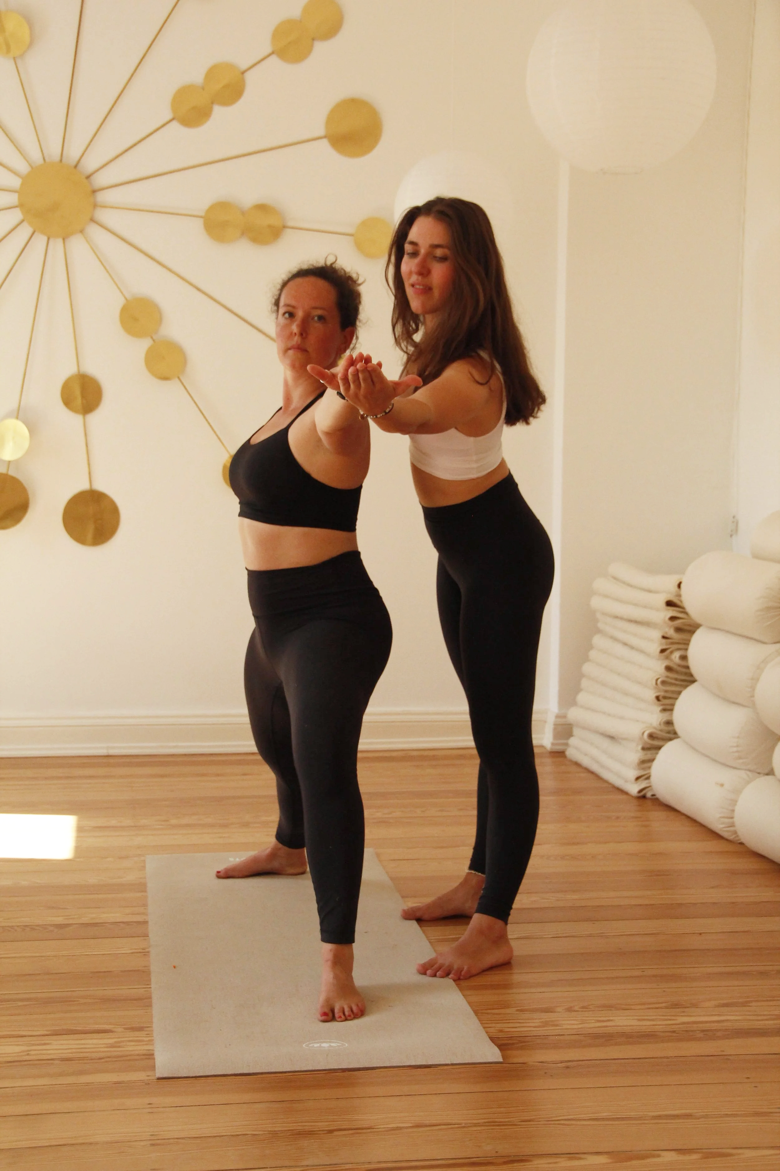 Zwei Frauen machen Yoga auf einem Yogamatte in einem Raum mit Holzfußboden und dekorativer Wandgestaltung.