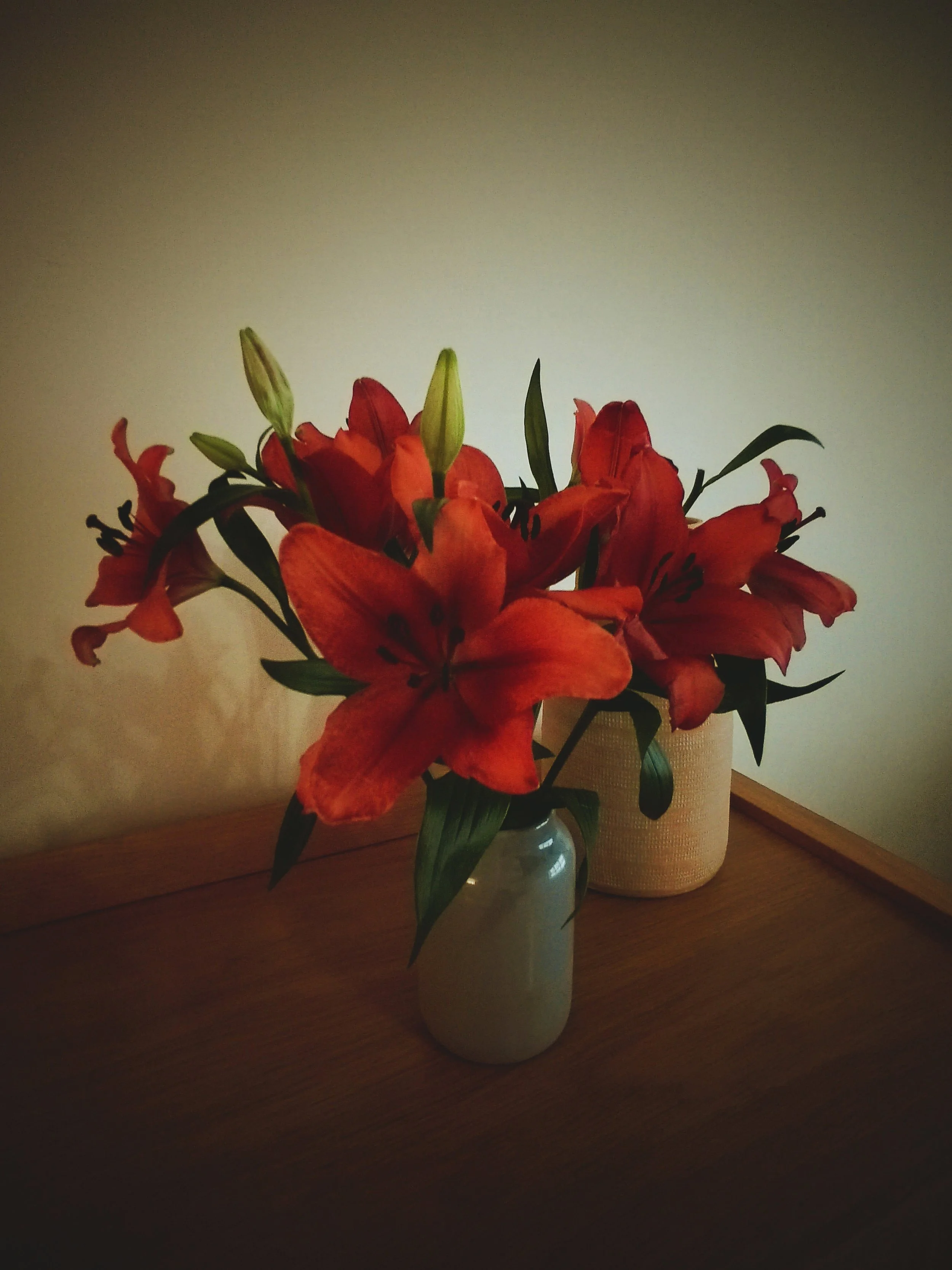 A bouquet of red lilies in a small gray vase on a wooden table, with a larger beigeish pot in the background.